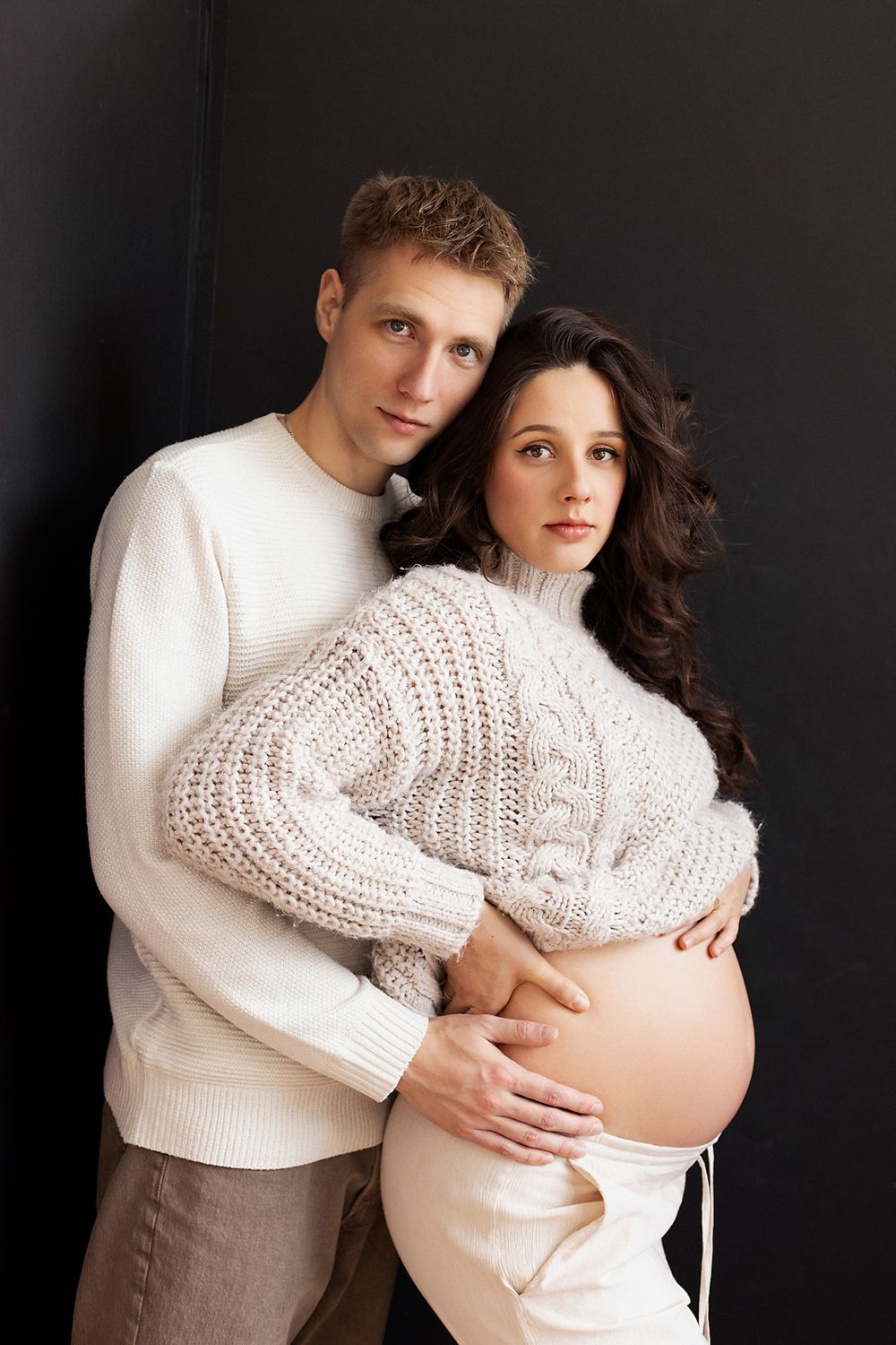 Couple in white knit sweaters embrace during maternity photo shoot against dark background.