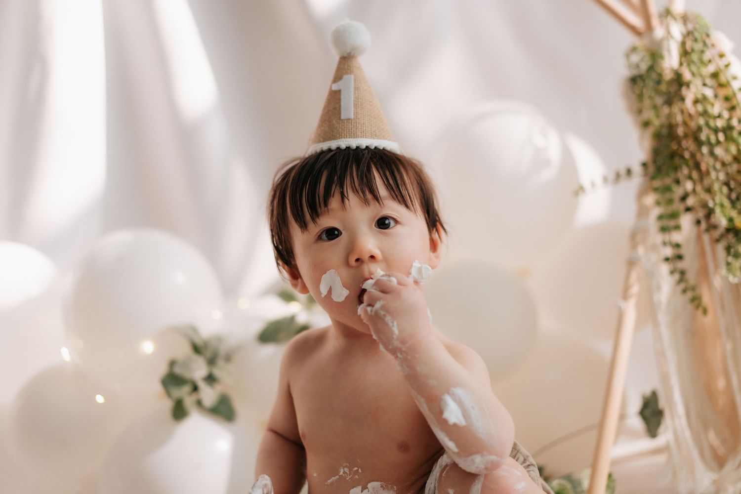 A baby sits in a white chair surrounded by floral decorations and balloons at a birthday celebration.