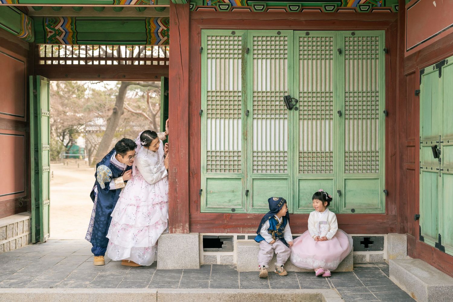 People in traditional Korean hanbok clothing pose in front of a historic palace building with green wooden doors.