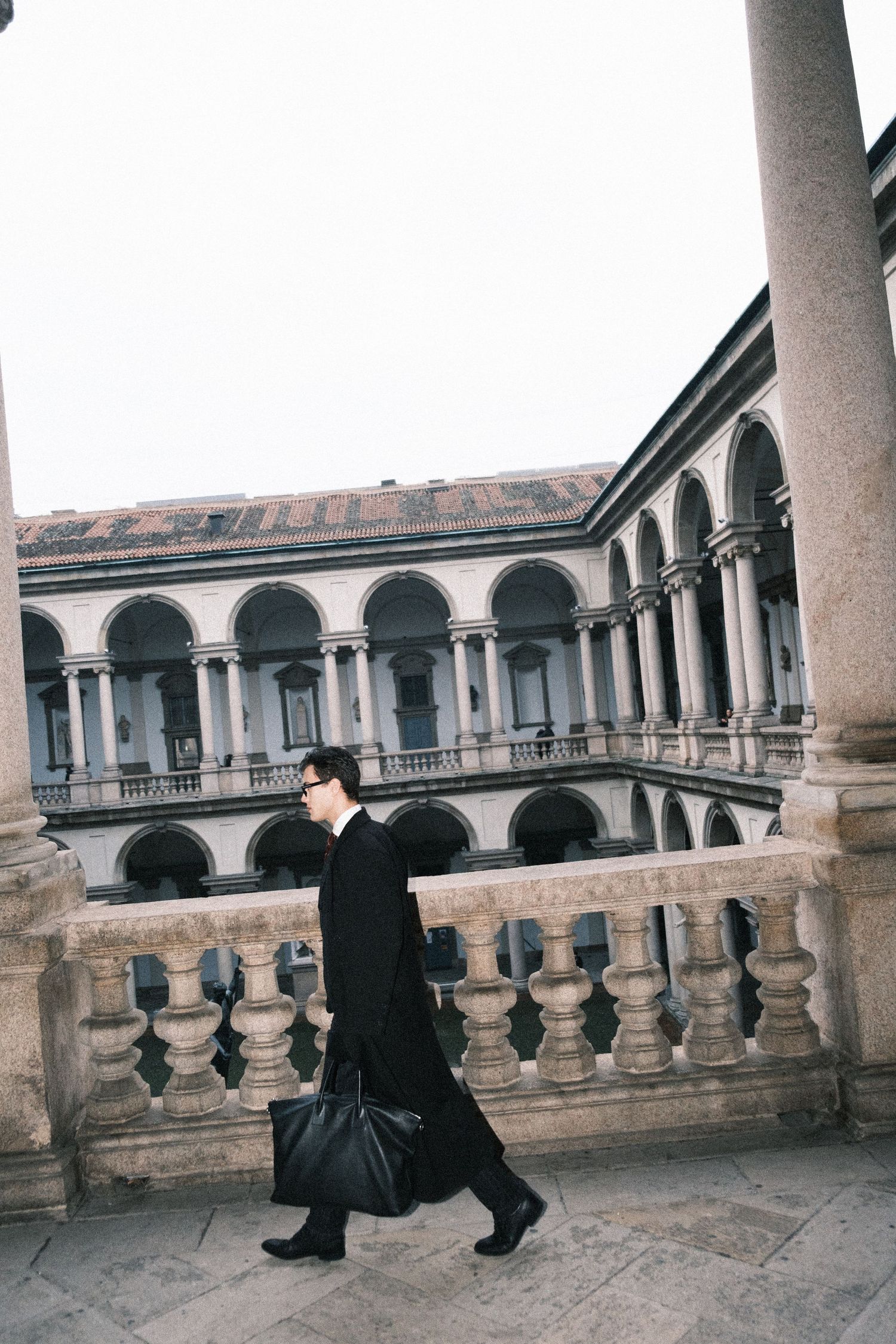 A person in dark clothing walks along a historic stone colonnade with arched architecture and balustrades.