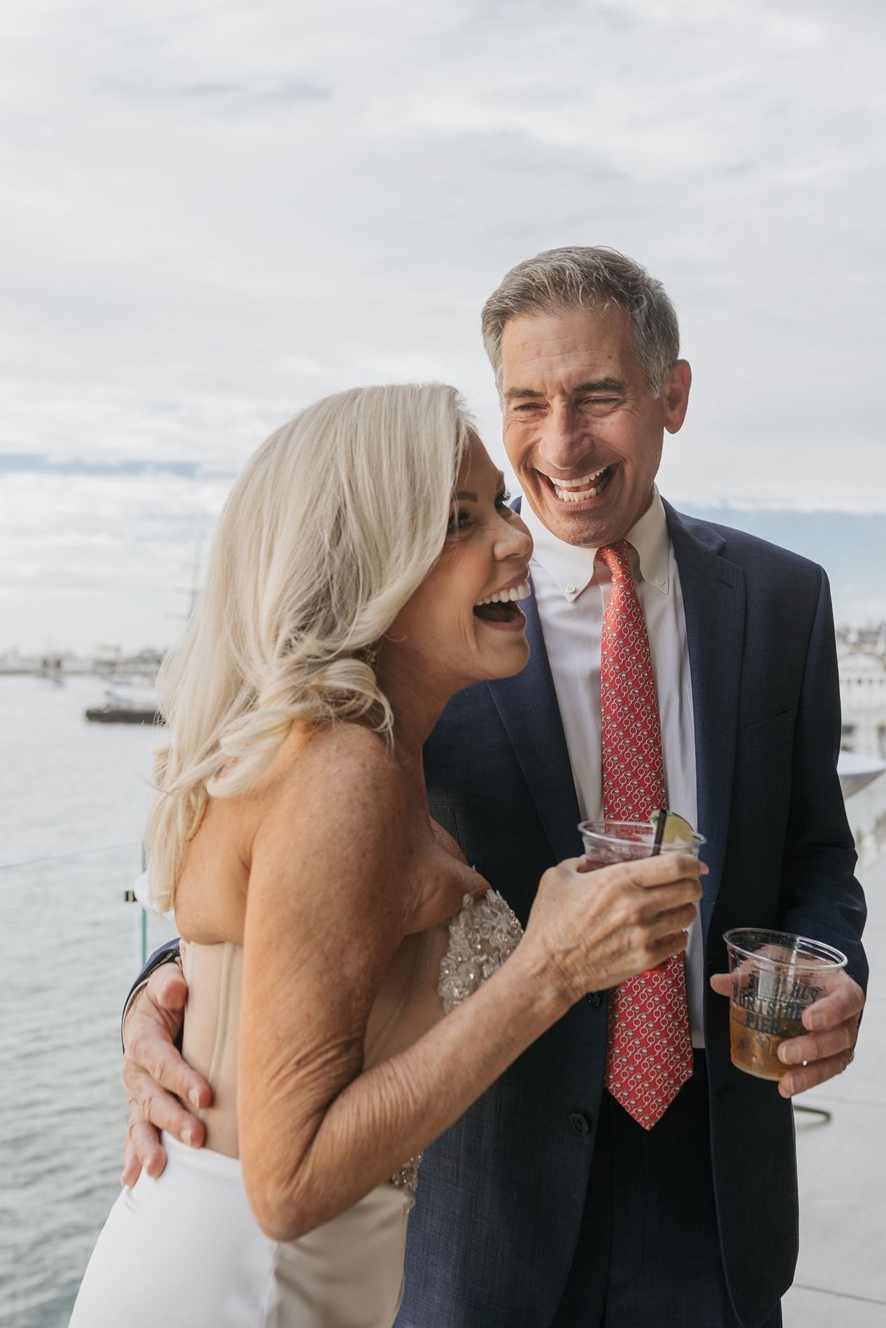 Happy couple laughing together and holding drinks at waterfront wedding celebration.