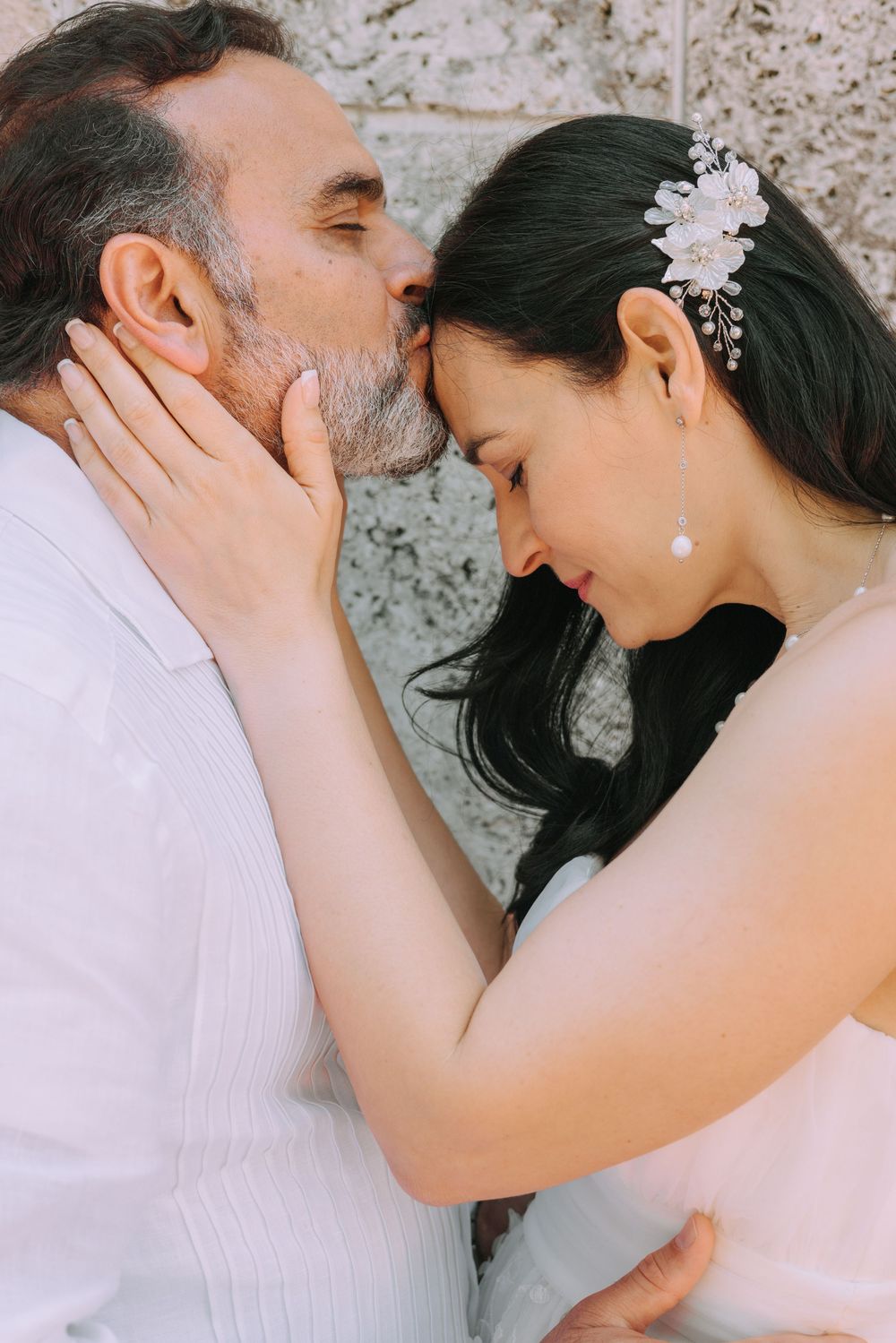 A tender moment between a bride with a floral hair accessory and someone leaning in close together against a textured wall.