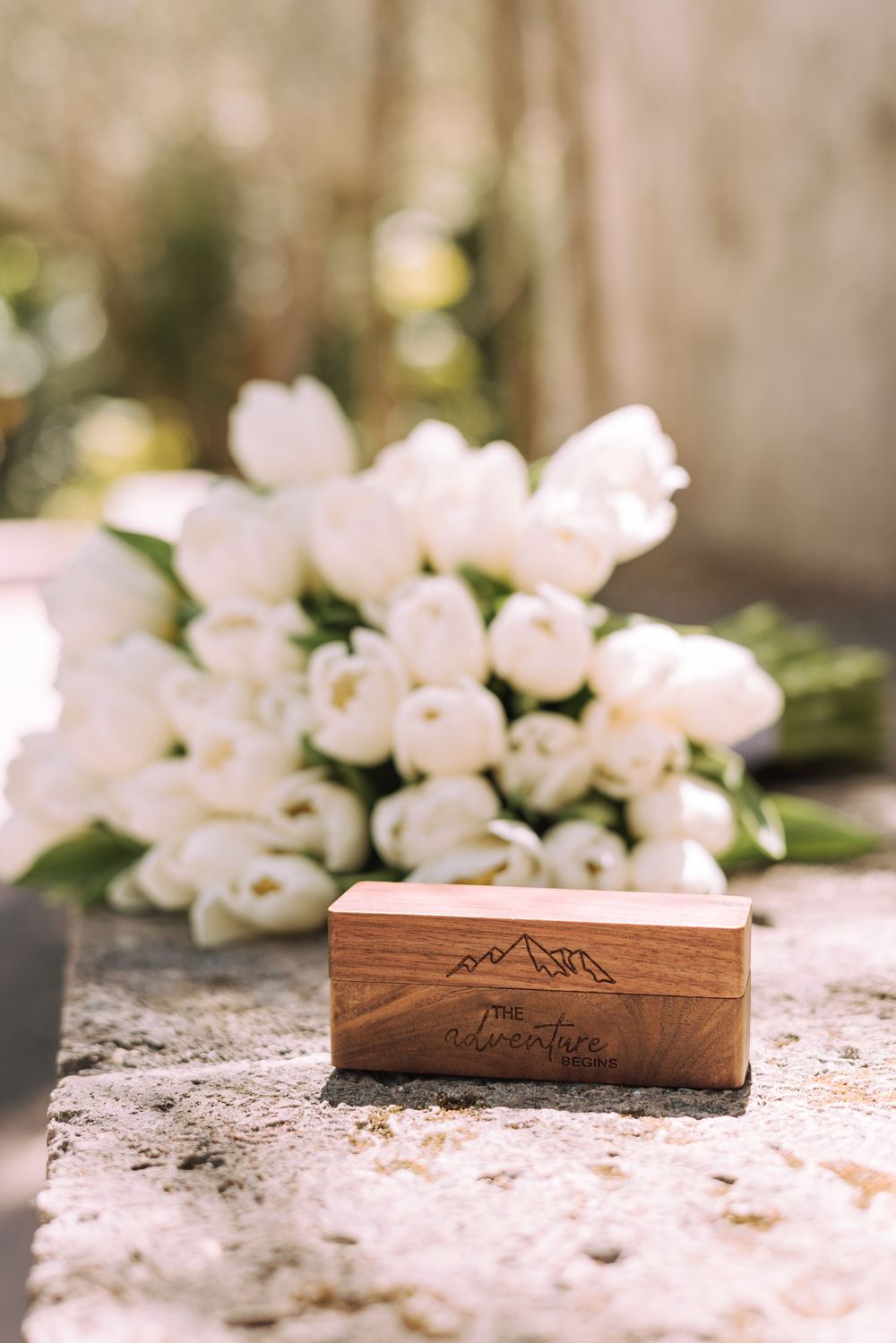 A wooden box with mountain engravings sits on a stone surface with delicate white flowers in the background.