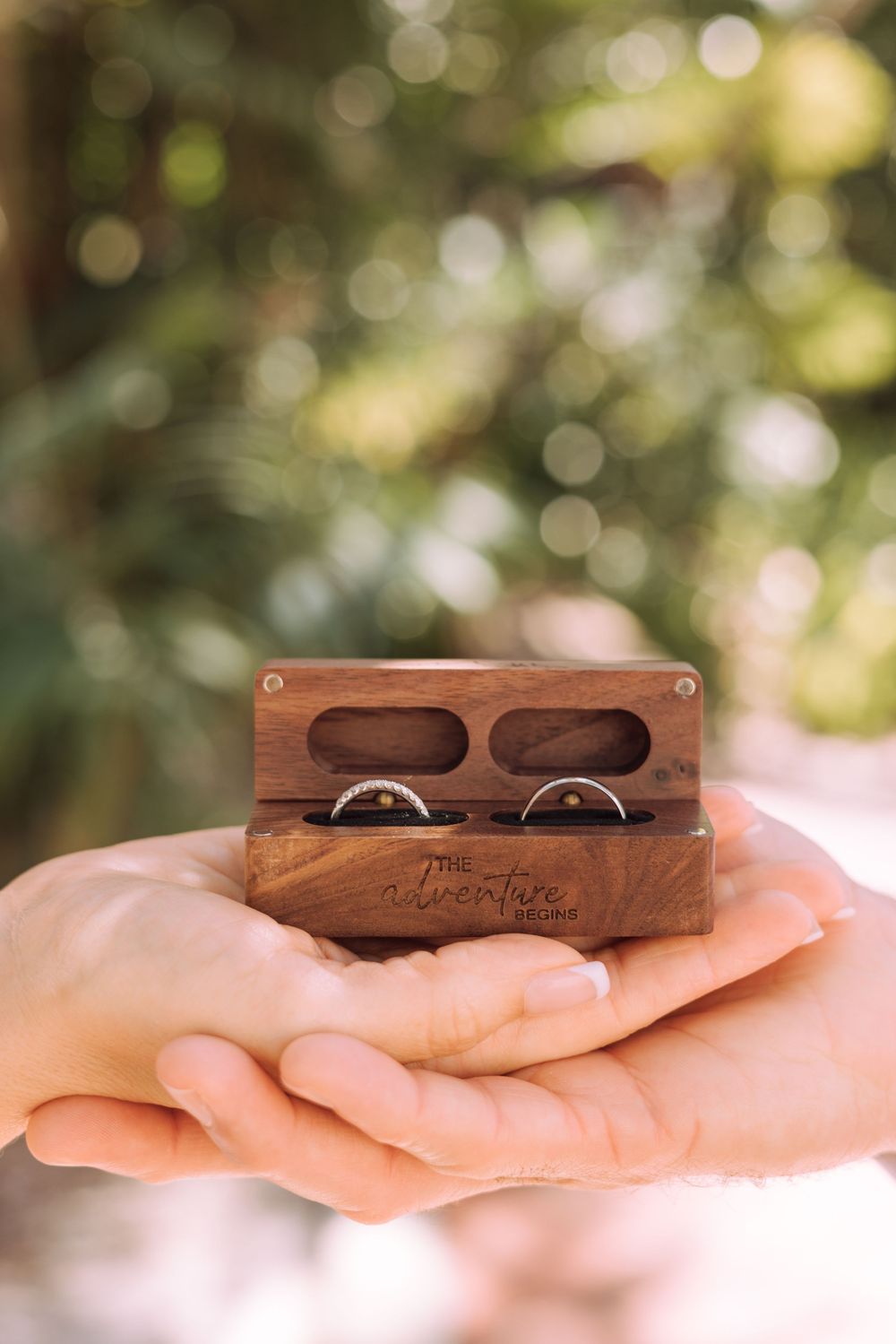 A wooden ring box with wedding bands is held in open palms against a soft blurred natural background.