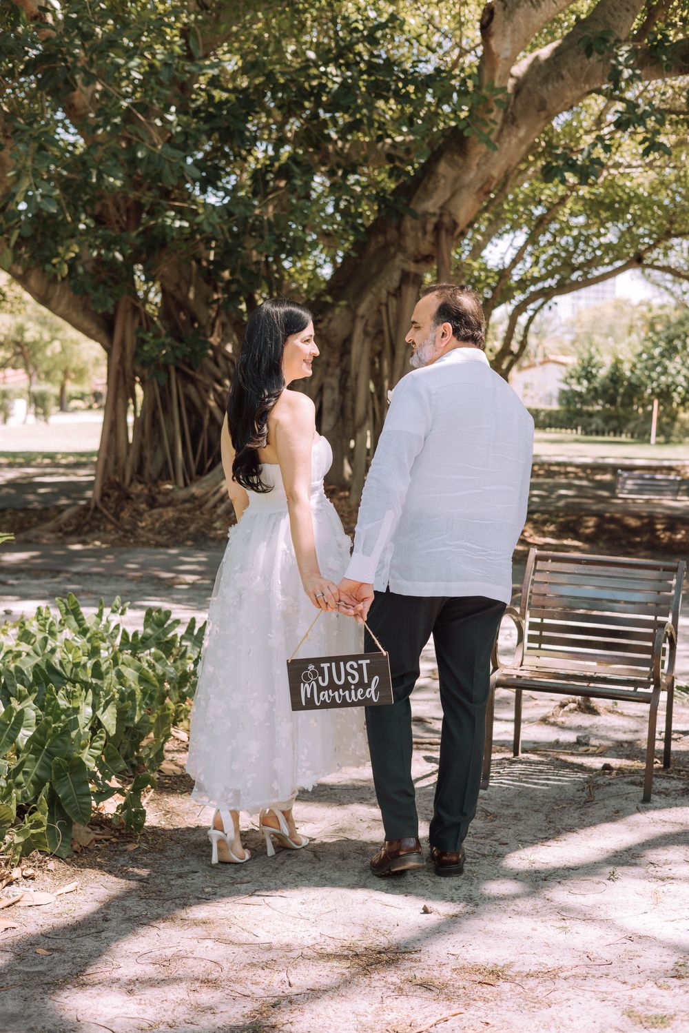 A couple in wedding attire sharing romantic moments while walking together under shady trees in a park setting.