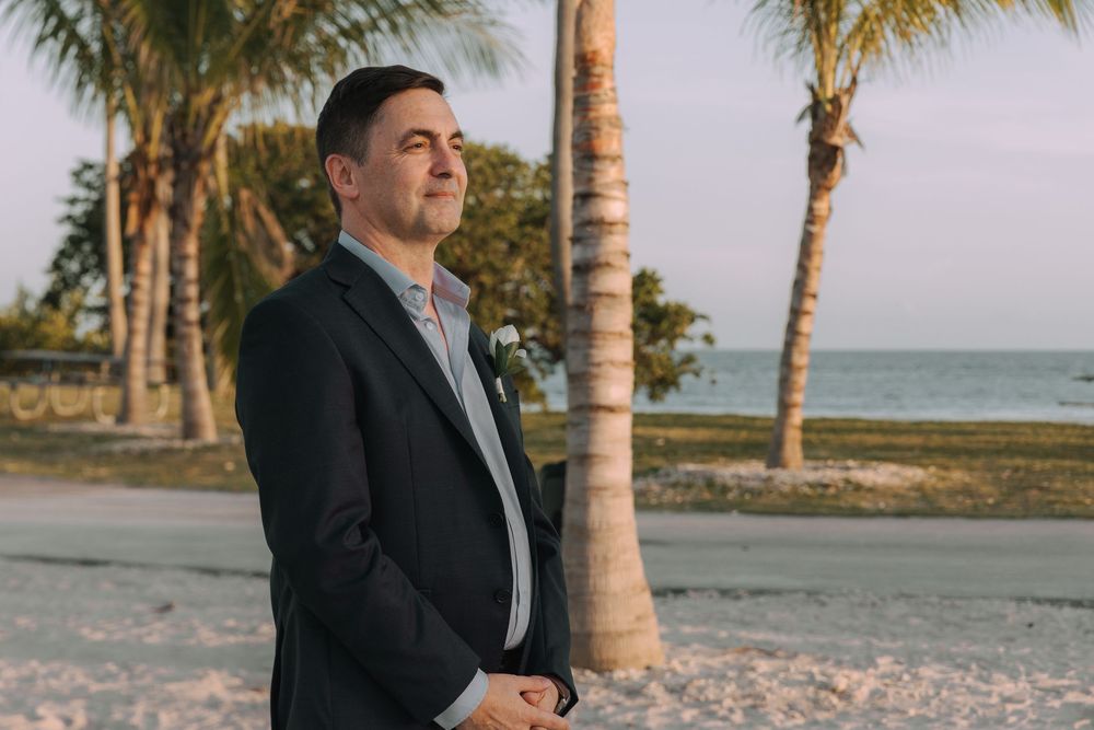 A person in a dark suit stands thoughtfully on a beach with palm trees and ocean during golden hour.