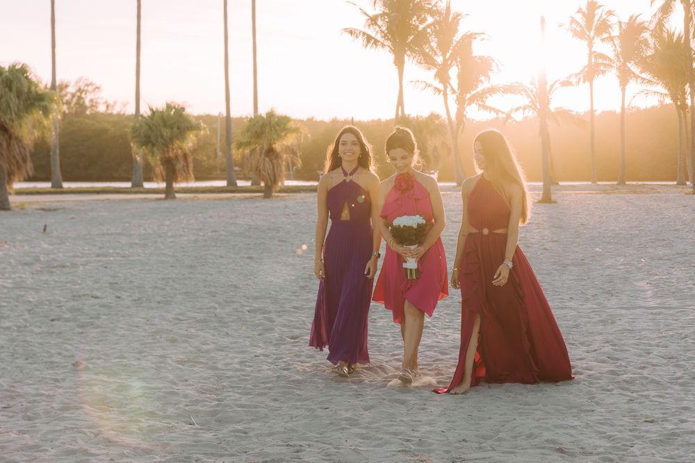 Three figures in flowing dresses pose together on a sandy beach during golden hour sunset.
