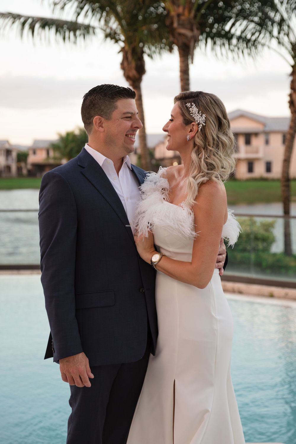 Romantic couple in formal wedding attire embraces by waterside during golden hour at luxurious venue.