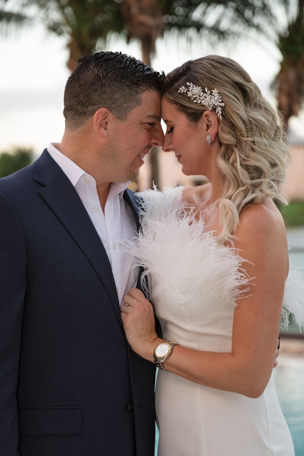 Romantic wedding couple shares tender moments in white attire against tropical palm background at sunset.