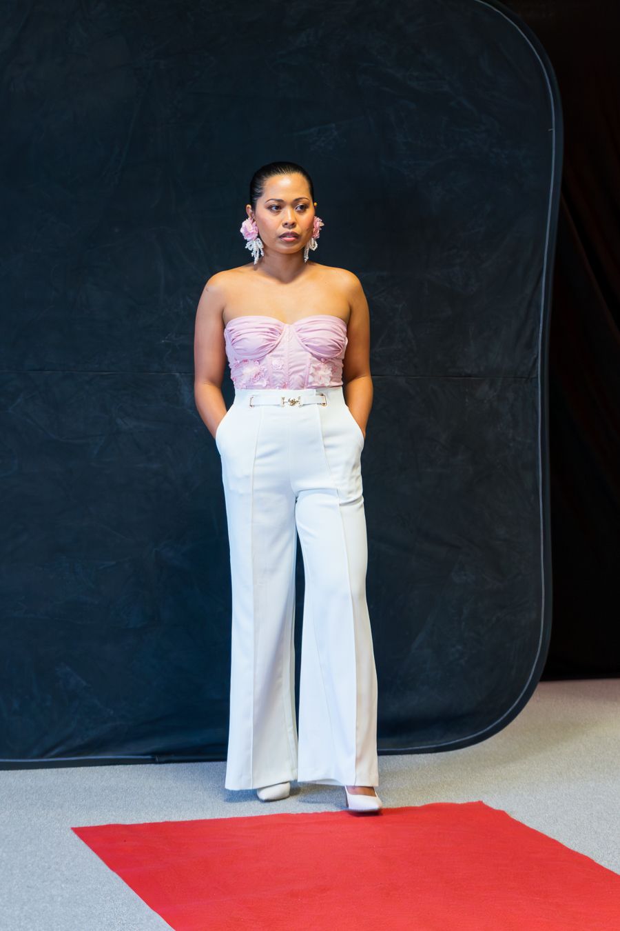 Model wearing a flowing white strapless jumpsuit poses against dark backdrop on red carpet.