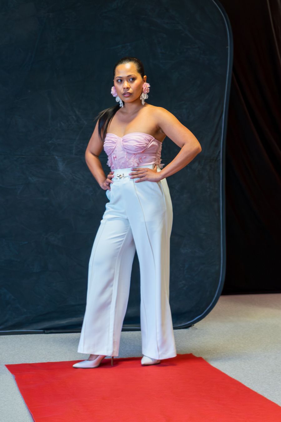 A person wearing a white jumpsuit and pink top poses on a red carpet against a dark backdrop.