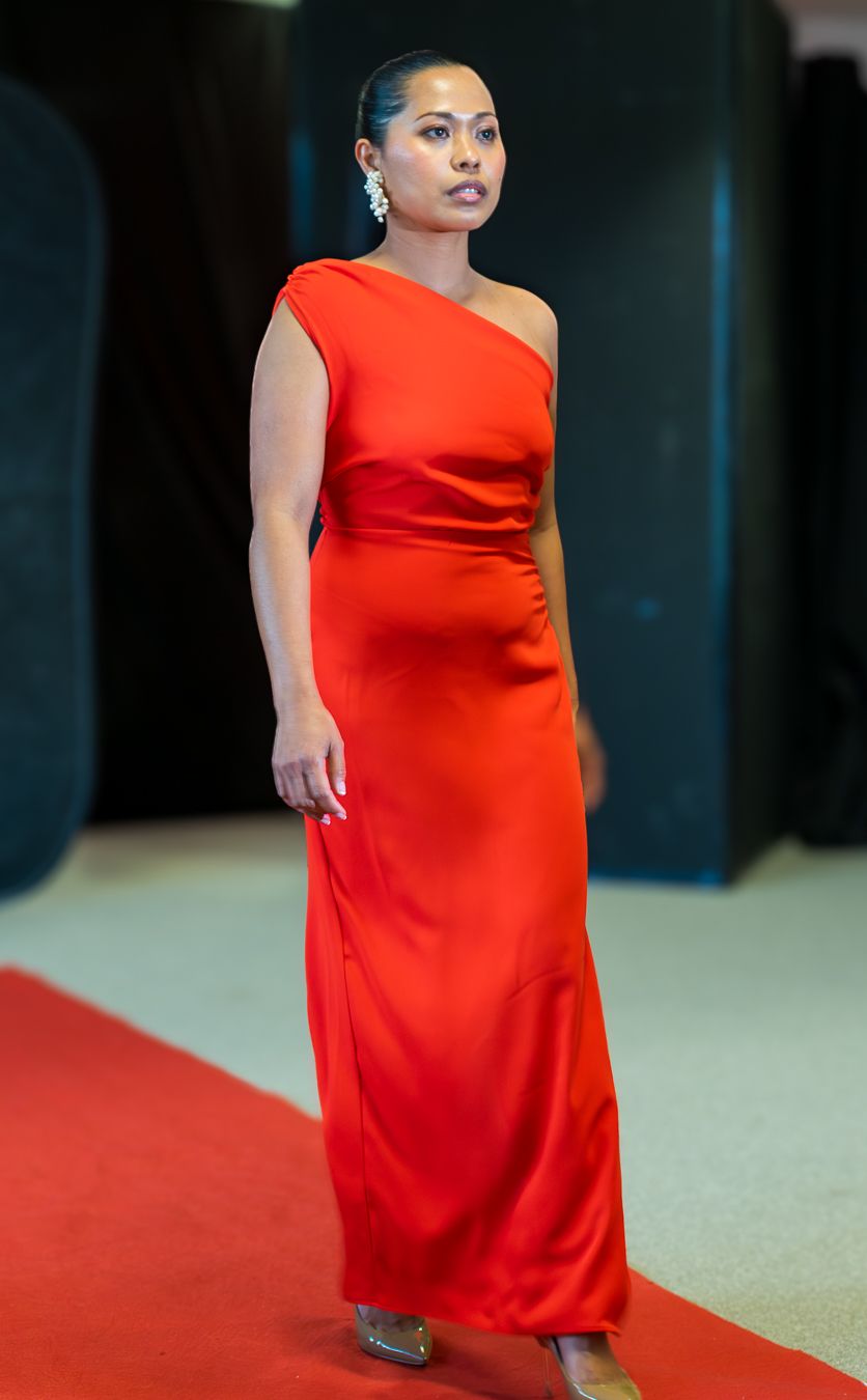 Model in elegant red one-shoulder evening gown poses on red carpet during fashion event.
