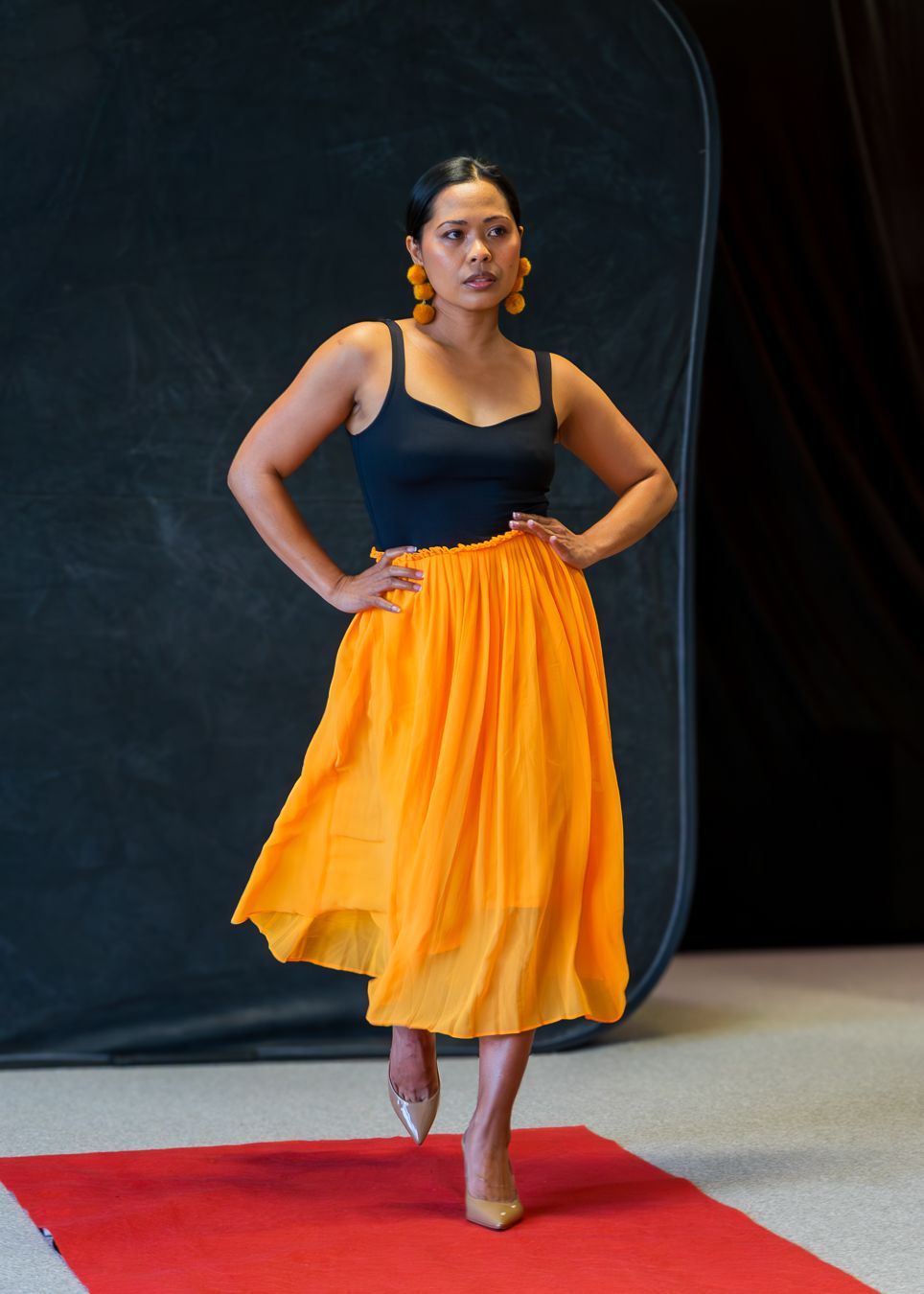 A dancer in a flowing orange dress performs a dynamic movement sequence on a red floor mat against a dark background.