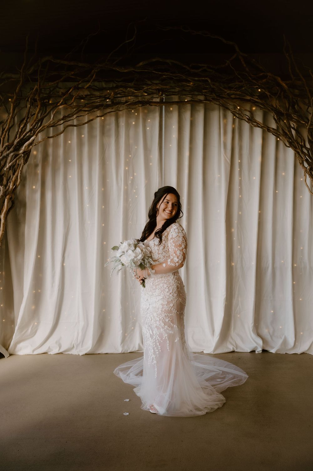 A bride in a lace long-sleeved wedding gown holds a white bouquet under a rustic arch with draped white curtains.