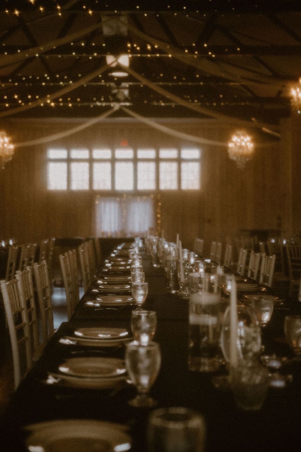 Long banquet table set for an elegant dinner under twinkling lights in a rustic reception venue with large windows.