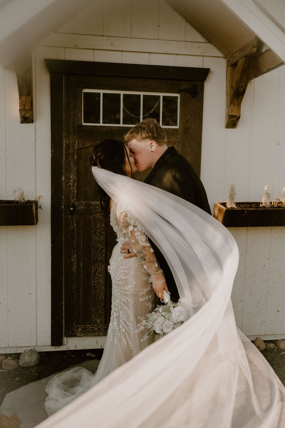 A flowing white wedding dress veil billows dramatically in the wind against a rustic building backdrop.