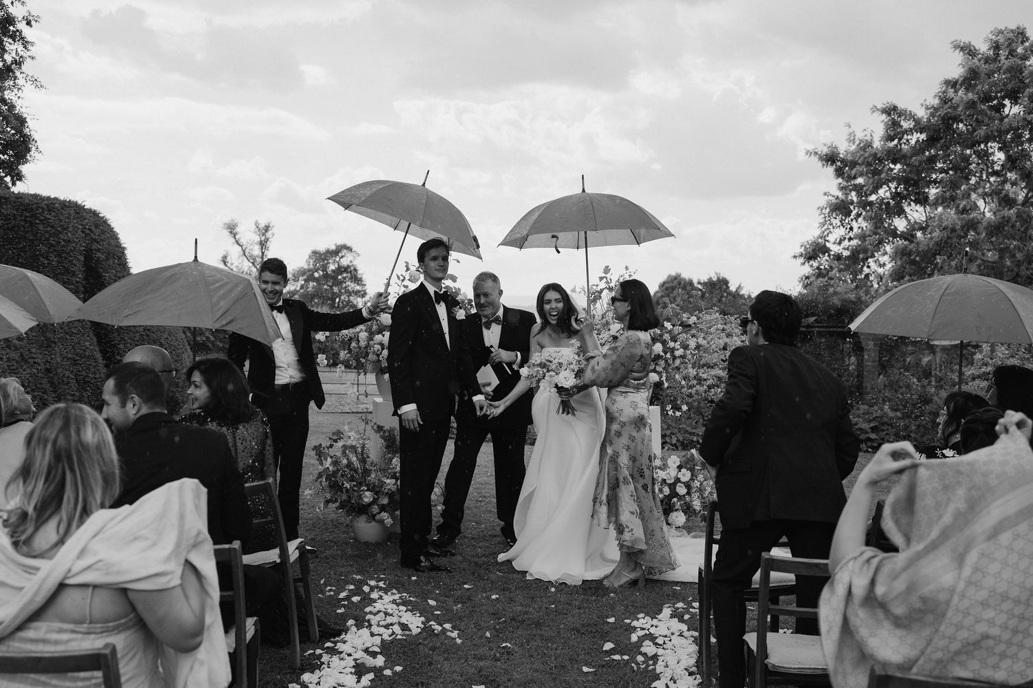 wedding guests, bride and groom sheltering underneath umbrellas during a rain shower during the ceremony at dewsall court in herefordshire