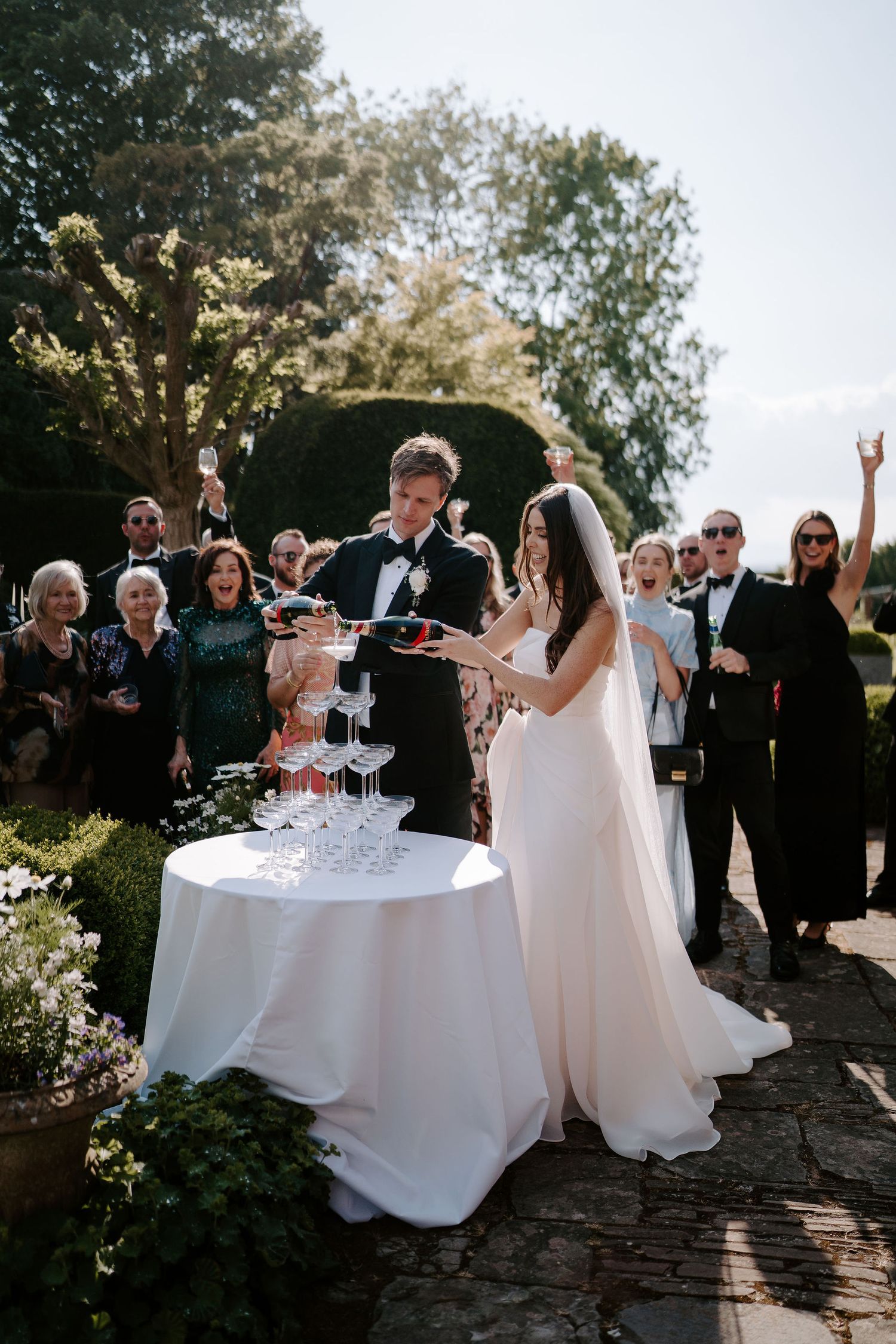 bride and groom pouring champagne into a tower of champagne glasses while their guests surround them cheering at dewsall court in hereford