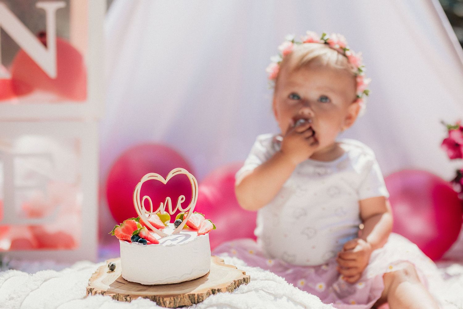 Bábätko s kvetinovou čelenkou pri narodeninovej torte zdobenej bobuľovým ovocím a srdiečkovým zápichom „One“. Žilina.