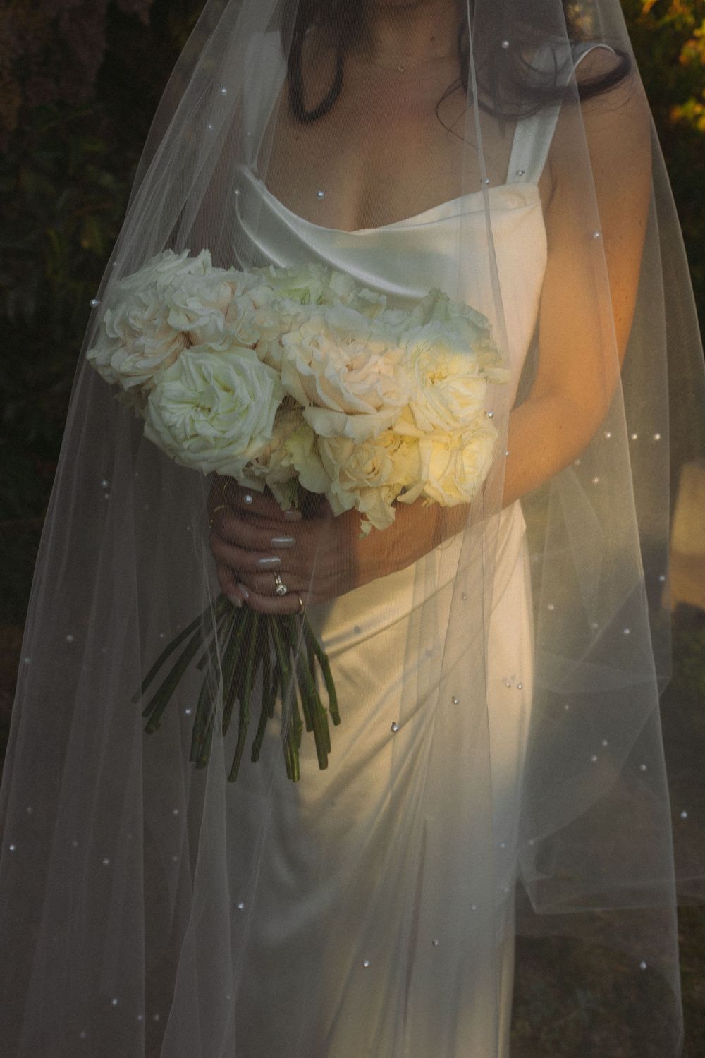 A bride in a flowing white dress holds an elegant bouquet of white roses under a delicate veil at sunset.