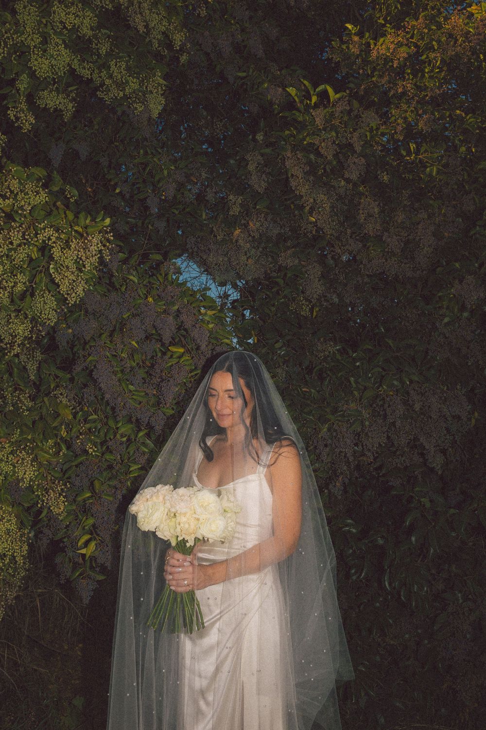 A bride in a flowing white gown and veil poses against a dark foliage background holding a white floral bouquet.