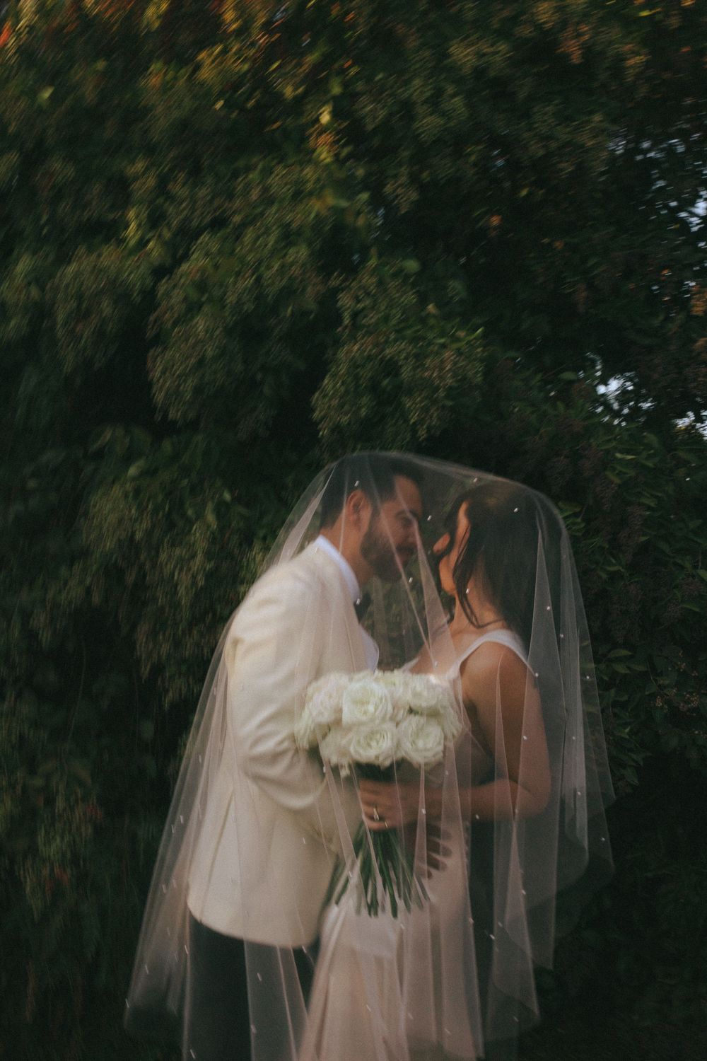 A romantic wedding couple shares a tender moment under a flowing veil in a garden setting.
