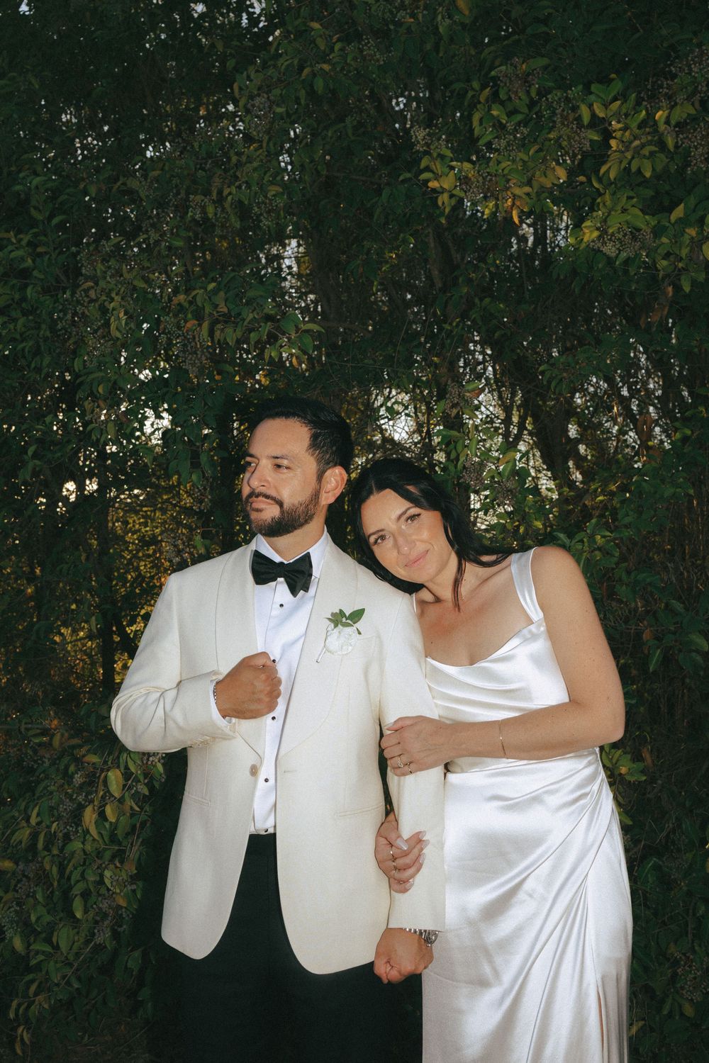 A couple in white wedding attire poses playfully in front of lush greenery outdoors.