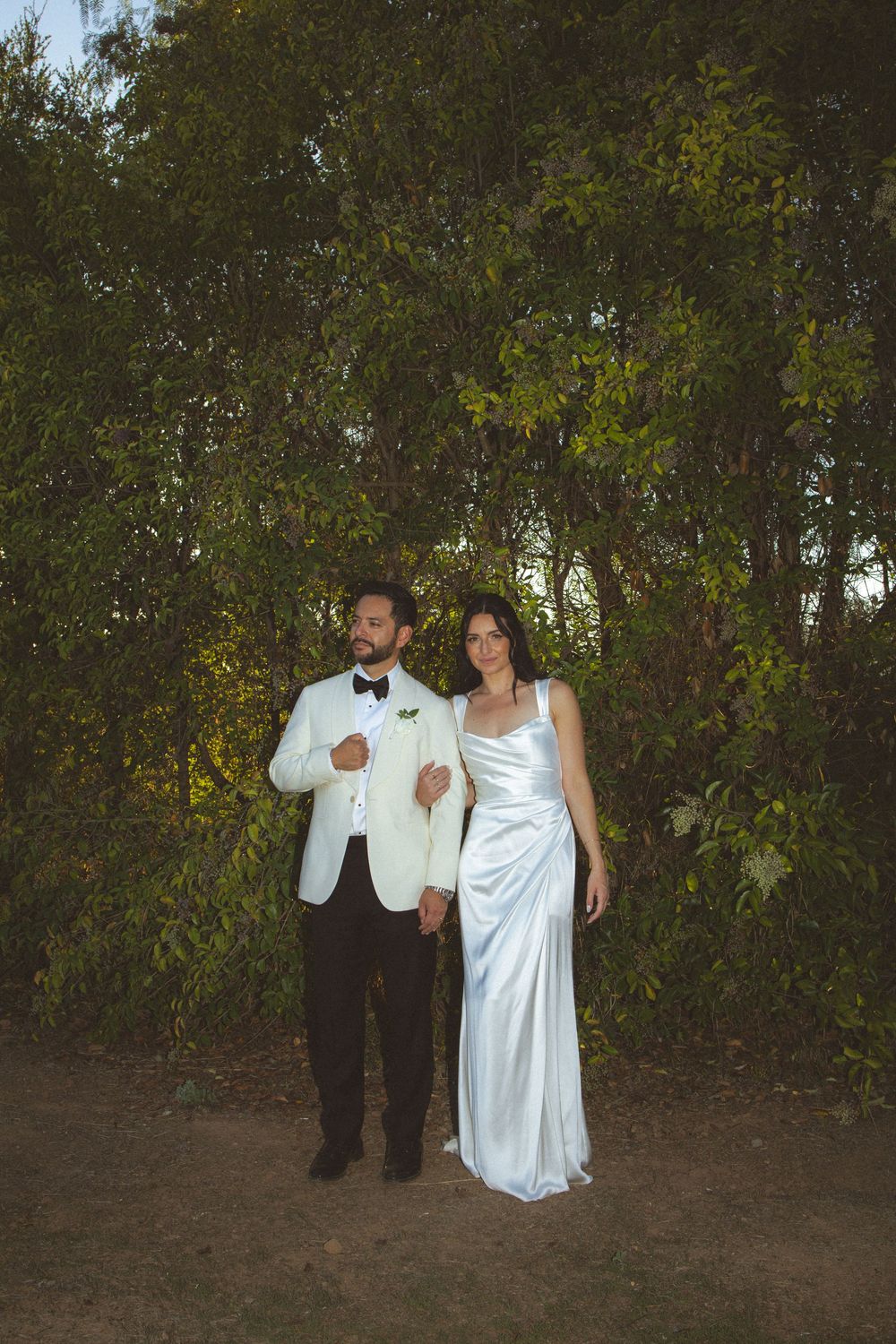 A couple in formal attire poses together in front of greenery during an outdoor evening event.