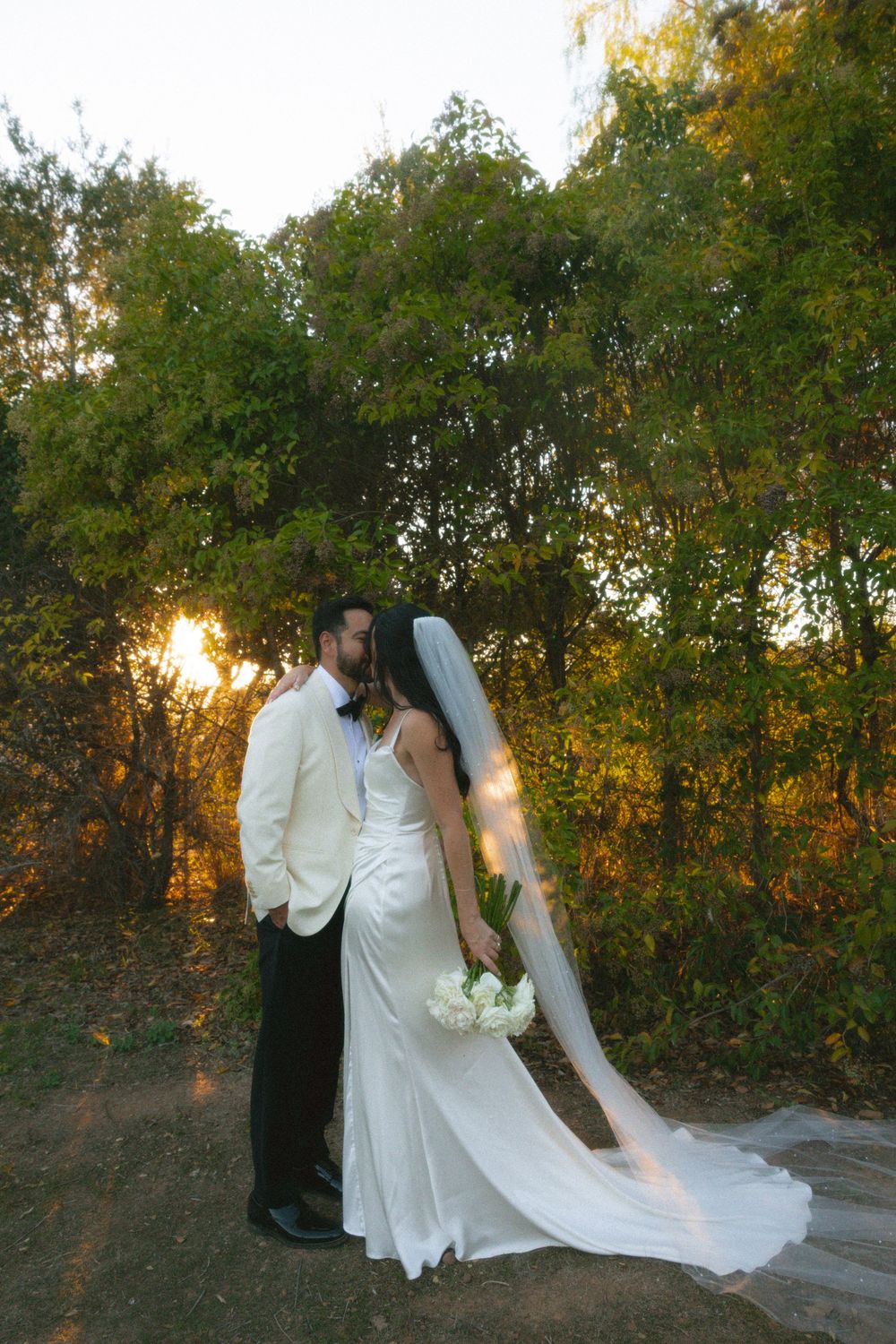 Newlyweds share an intimate moment at sunset among autumn trees in an outdoor garden setting.