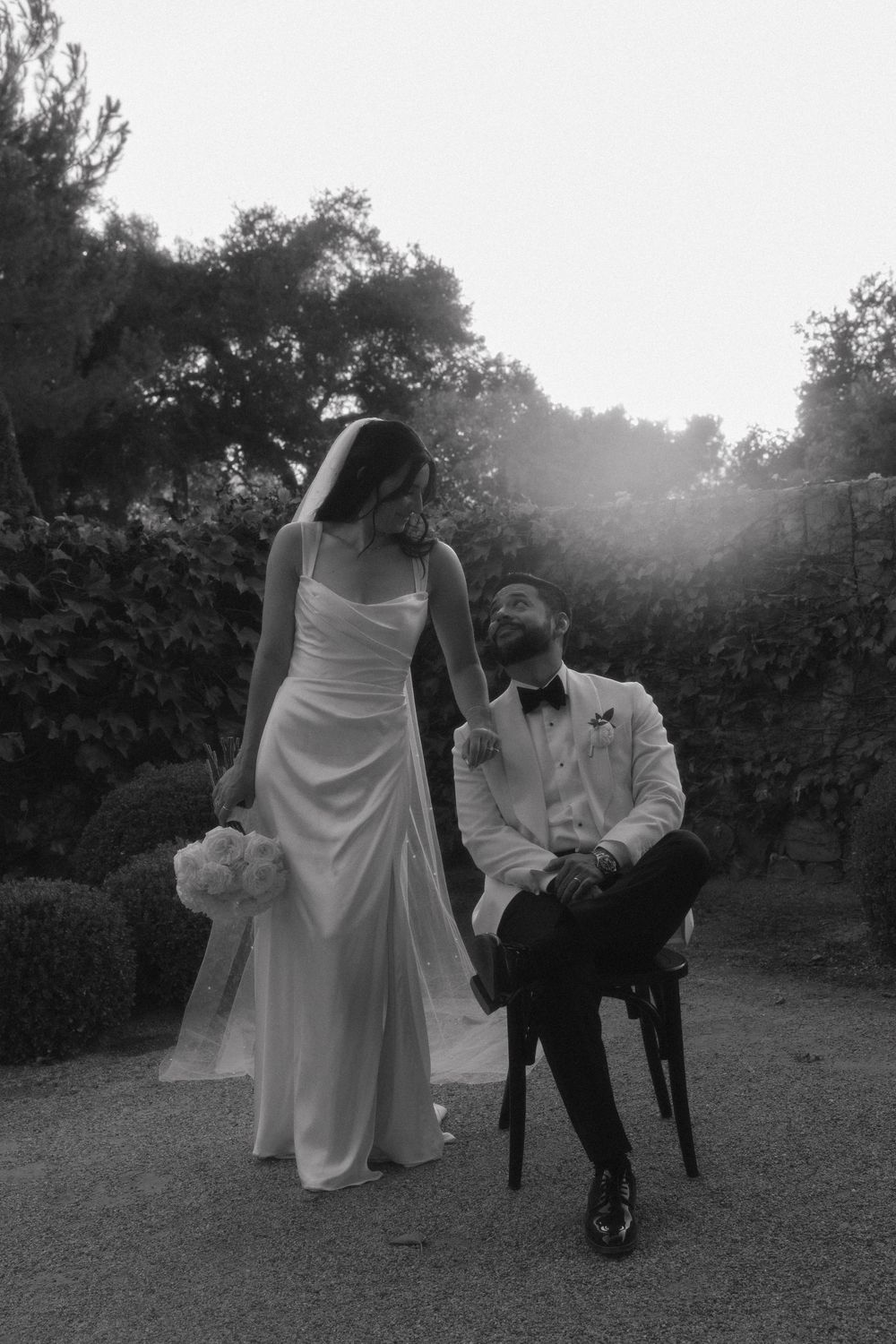 A black and white wedding photo of a couple outdoors, with the bride standing and groom seated on a chair.