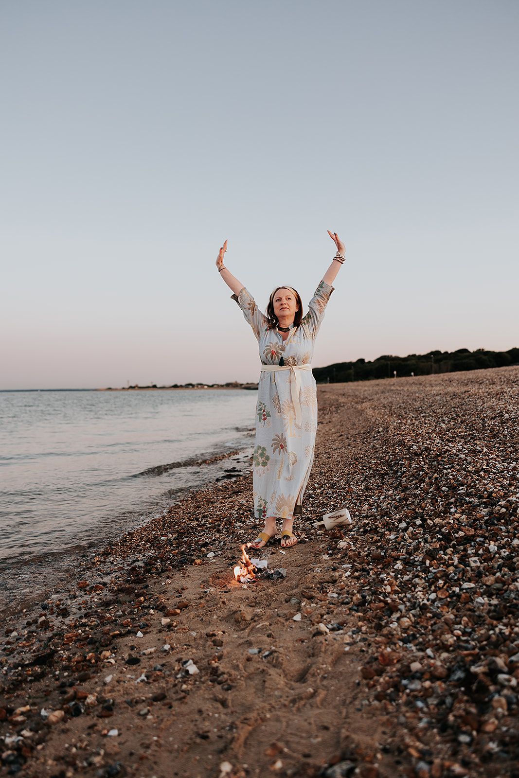 A figure in a flowing white dress jumps joyfully on a rocky beach at sunset with arms raised to the sky.
