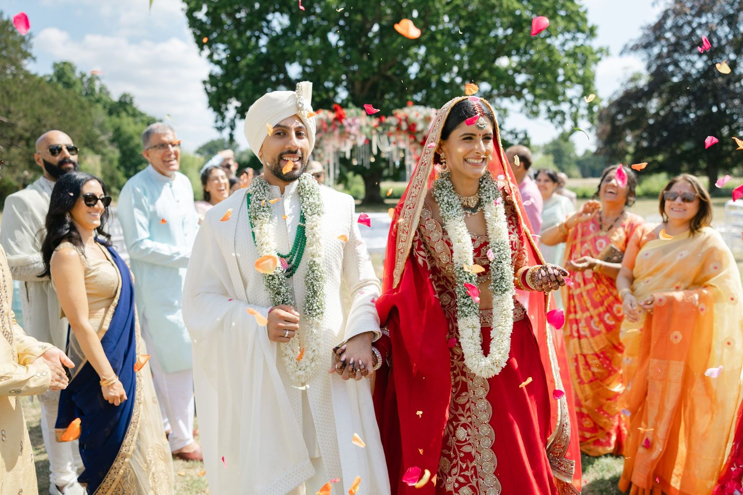 A radiant Hindu bride and groom walking through a colorful petal toss celebration during an outdoor wedding ceremony at Ditton Manor