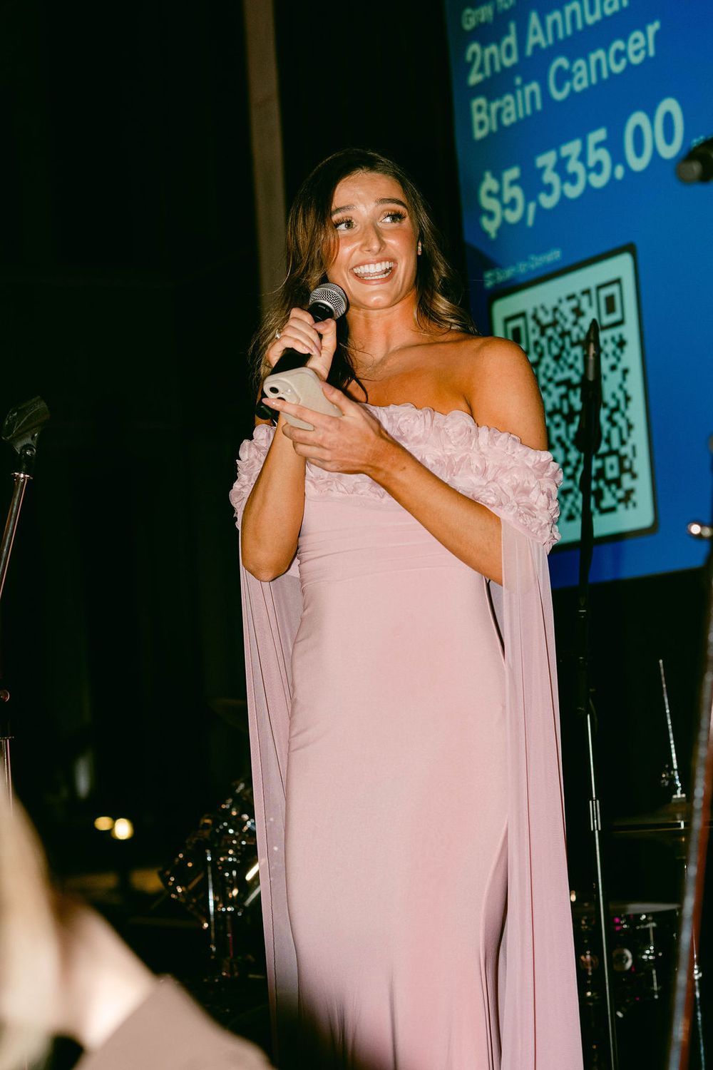 A person in a pink off-shoulder gown speaks into a microphone at a fundraising event for brain cancer.