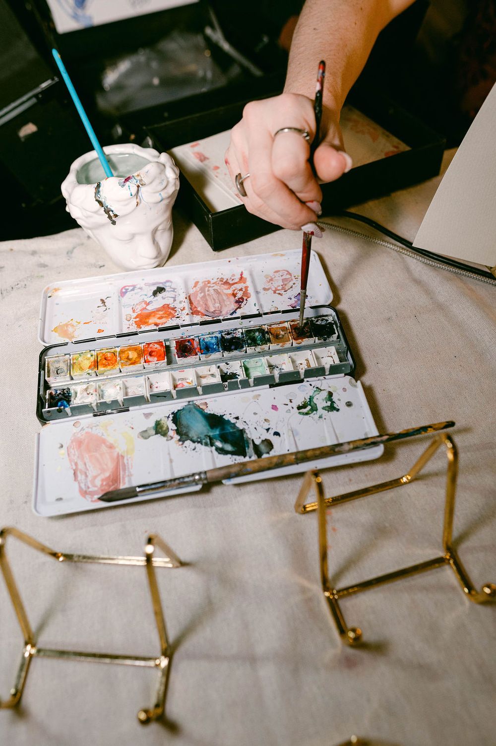Watercolor paint palette and brushes arranged on white paper with brass or gold colored metal objects.