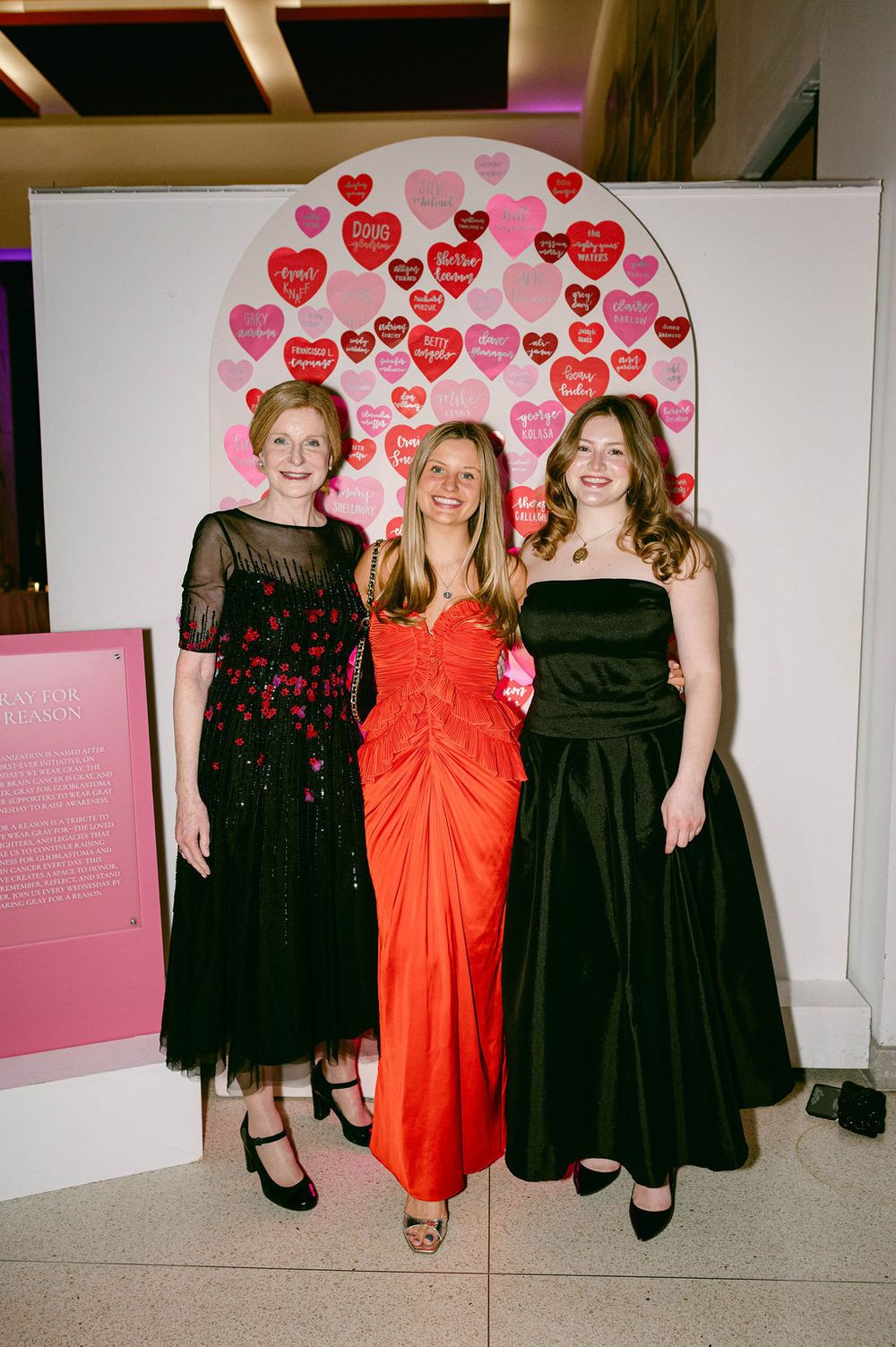 Three women pose together in formal dresses - one in red and two in black - against a wall with pink heart decorations.