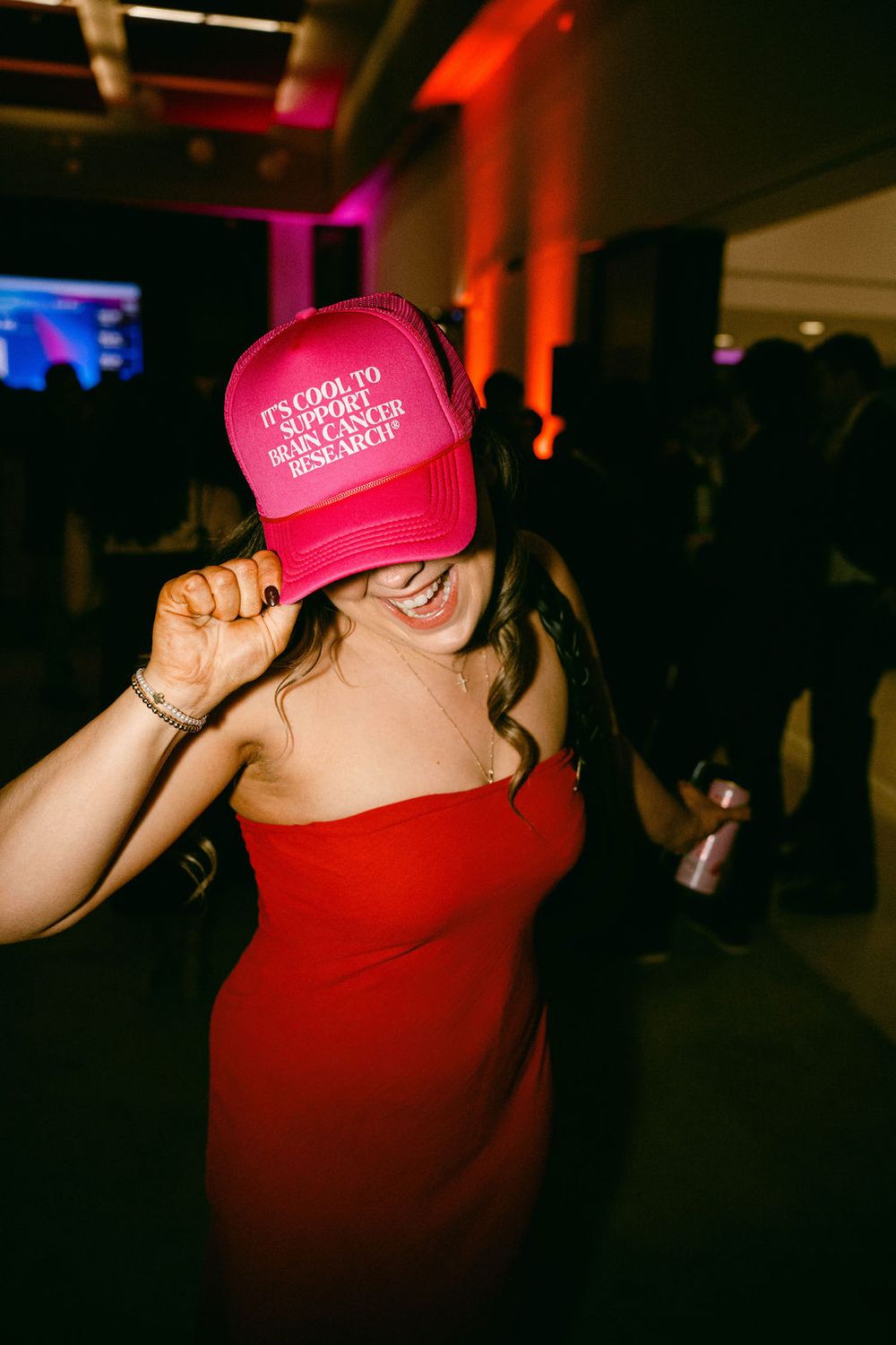 A person in a red top wearing a bright pink baseball cap dances in a dimly lit venue.