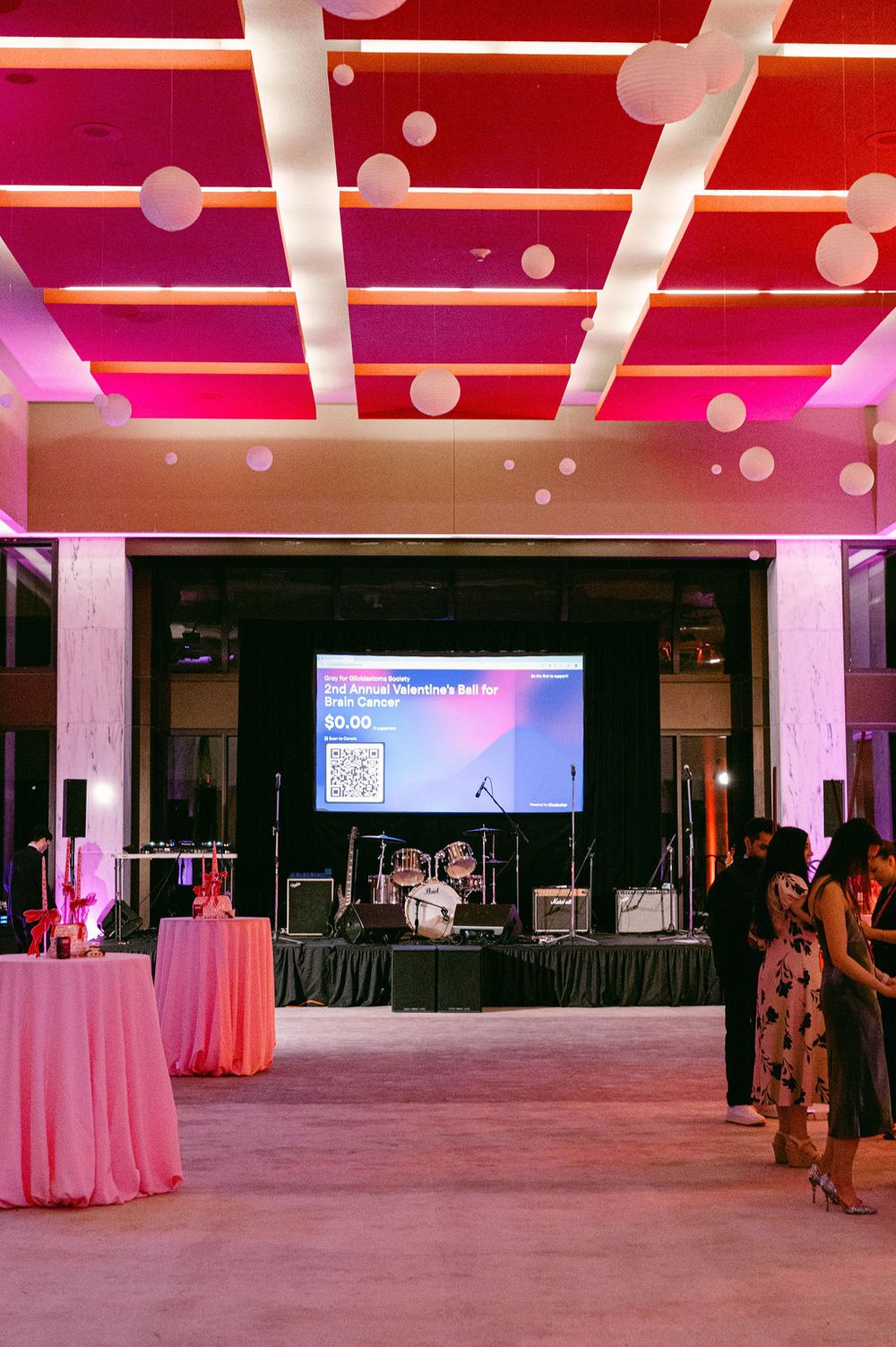 Large empty event space with red ceiling lights and white columns features scattered cocktail tables and projection screens.