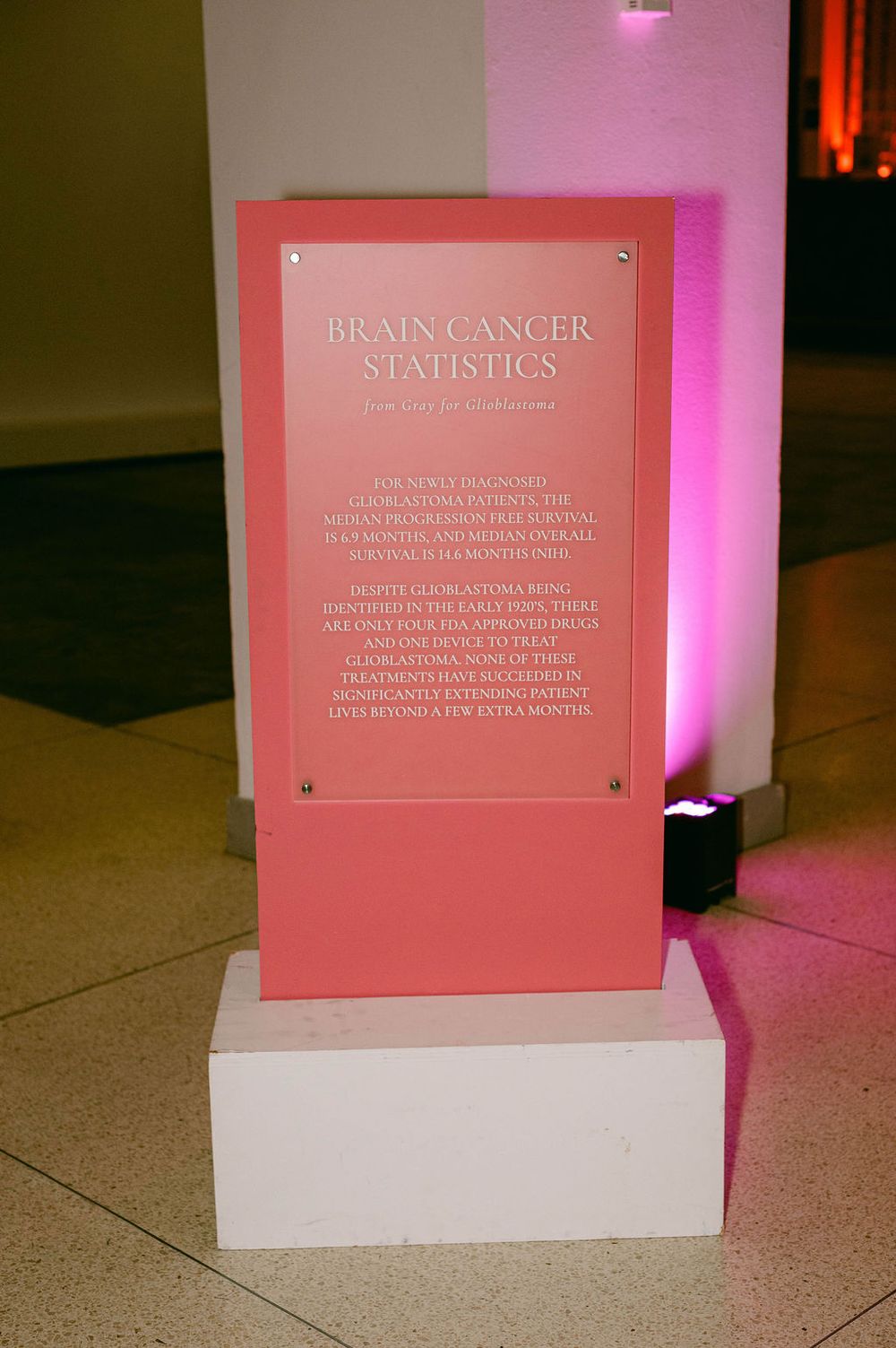 A pink informational plaque displaying brain cancer statistics stands on a white pedestal with pink lighting in the background.