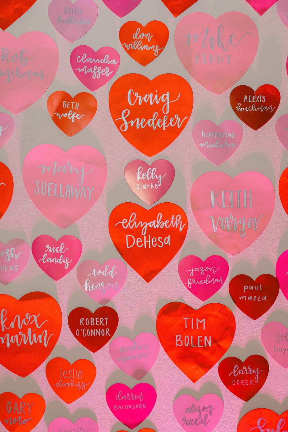 A colorful pattern of pink, red, and burgundy heart shapes with handwritten names creates a Valentine's Day themed background.