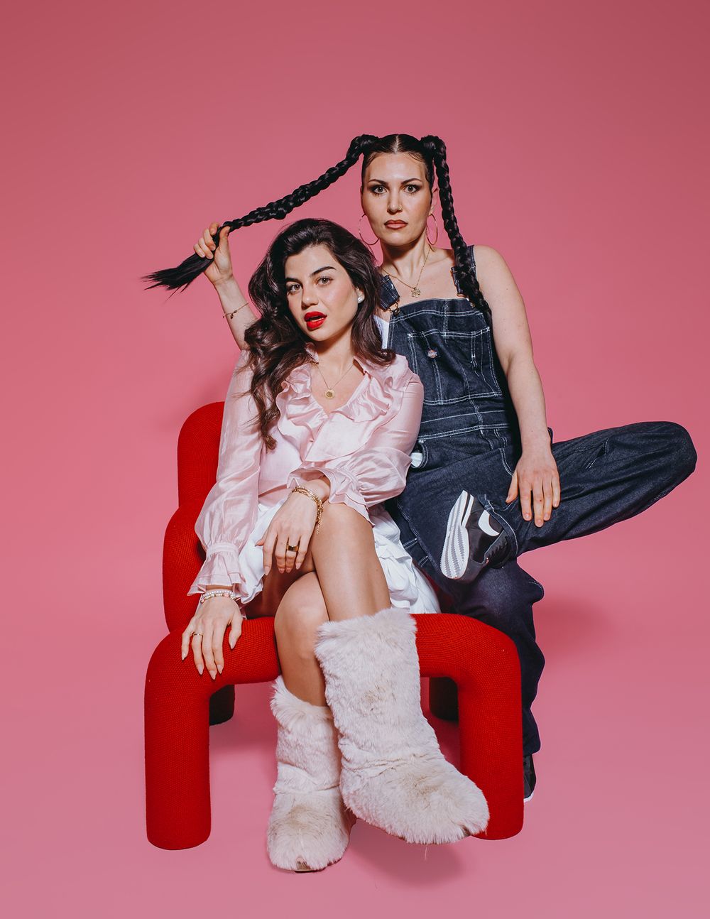 Couple in contrasting black and pink outfits pose dramatically on red velvet chair against pink studio backdrop.