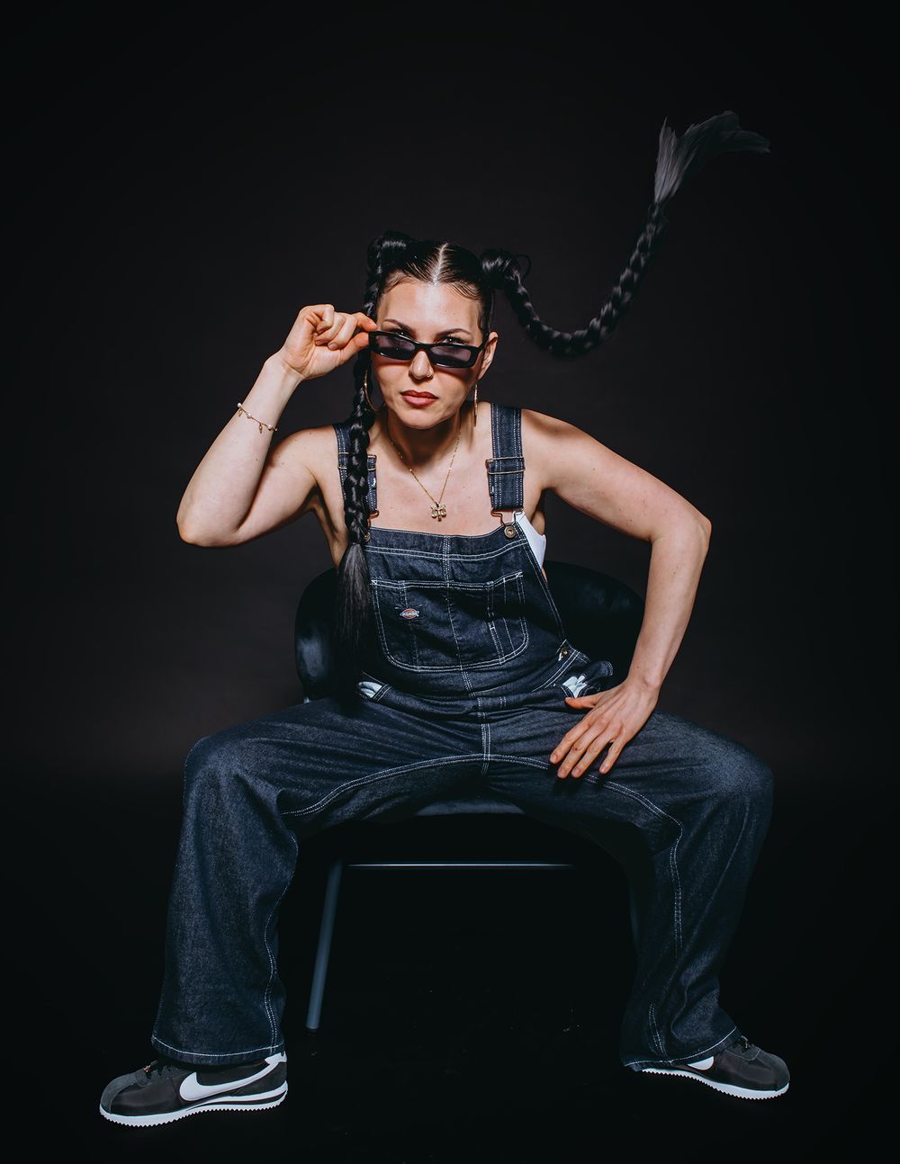 Person in denim overalls and sunglasses poses dramatically against dark background in artistic photo series.