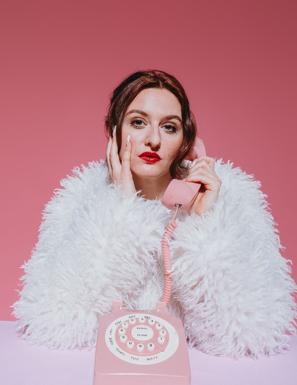 A model in a fluffy white feathered garment poses against a pink backdrop in a glamorous fashion portrait series.