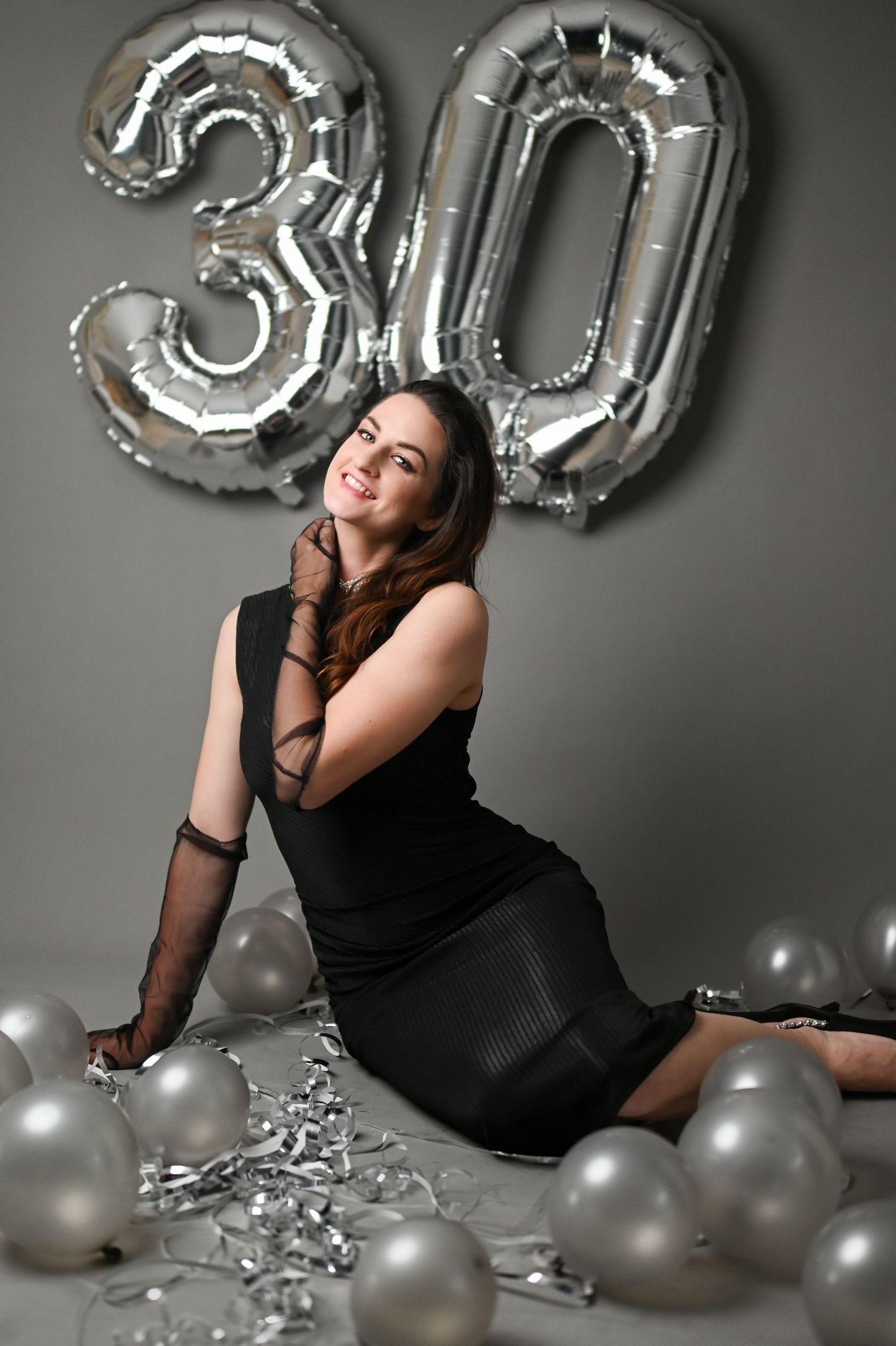 Celebratory 30th birthday photoshoot with silver number balloons and elegant black dress against grey backdrop.