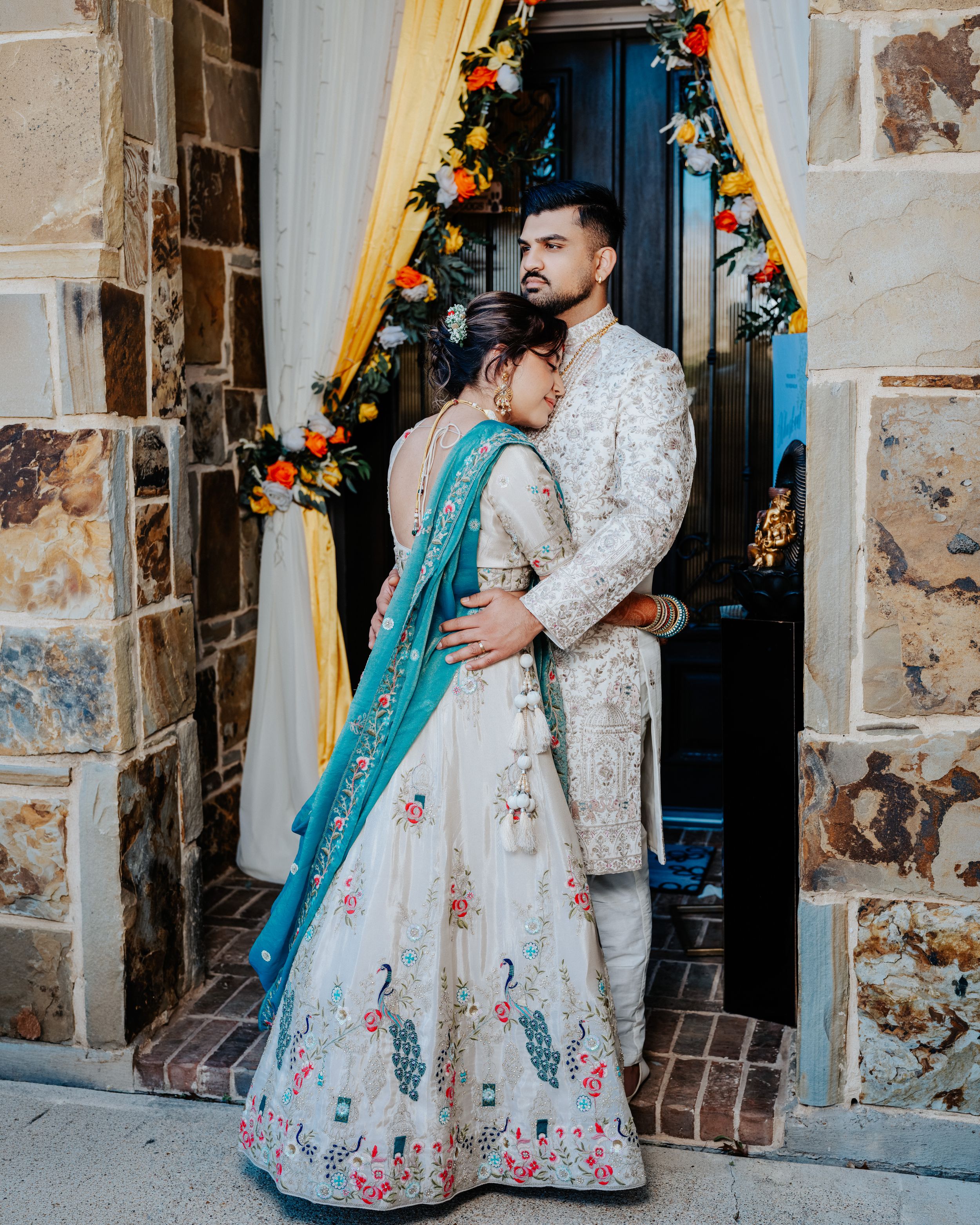 A couple in traditional South Asian wedding attire embrace in front of a decorated doorway with floral garlands.