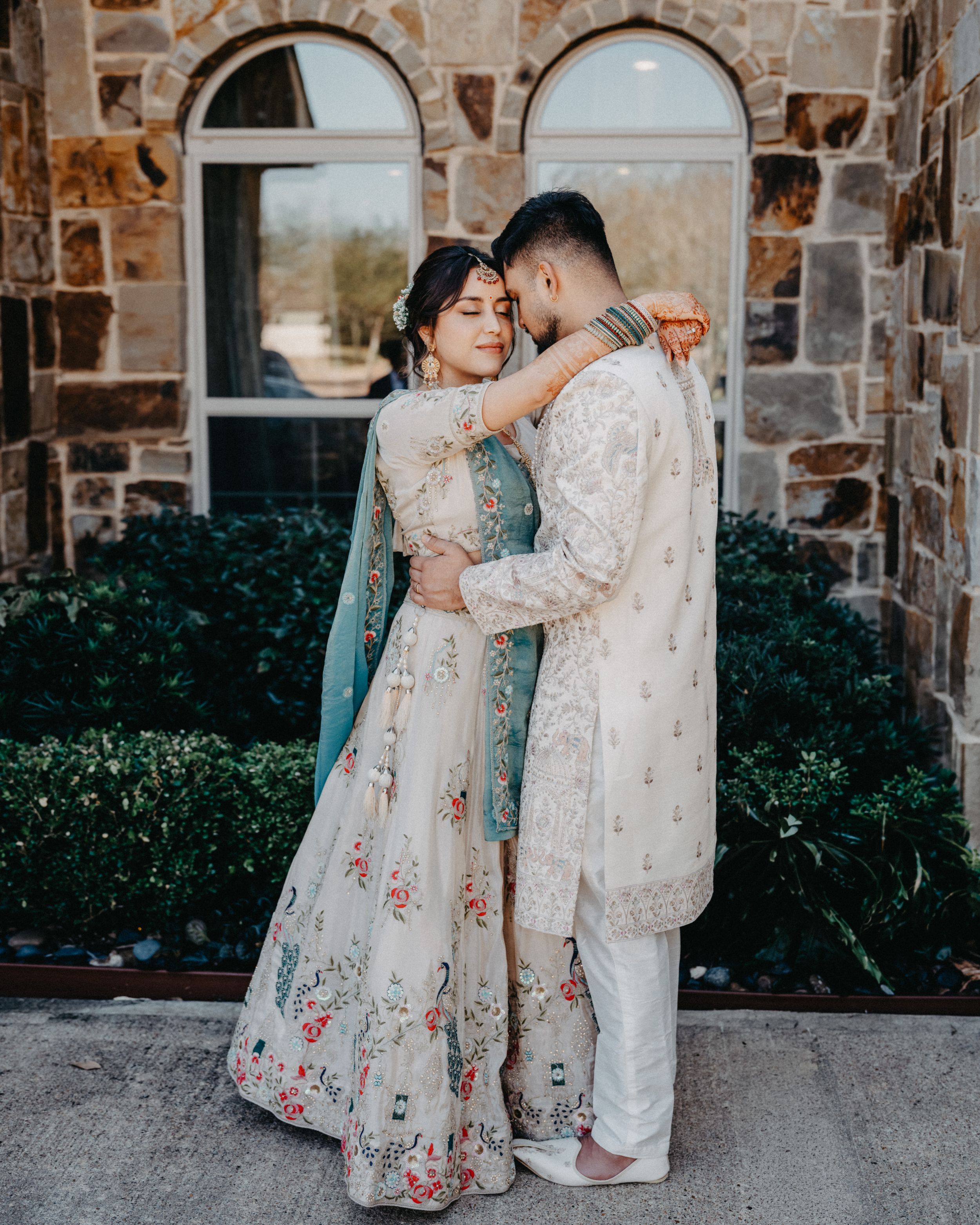 A couple in traditional South Asian wedding attire embraces in front of a stone building wall.