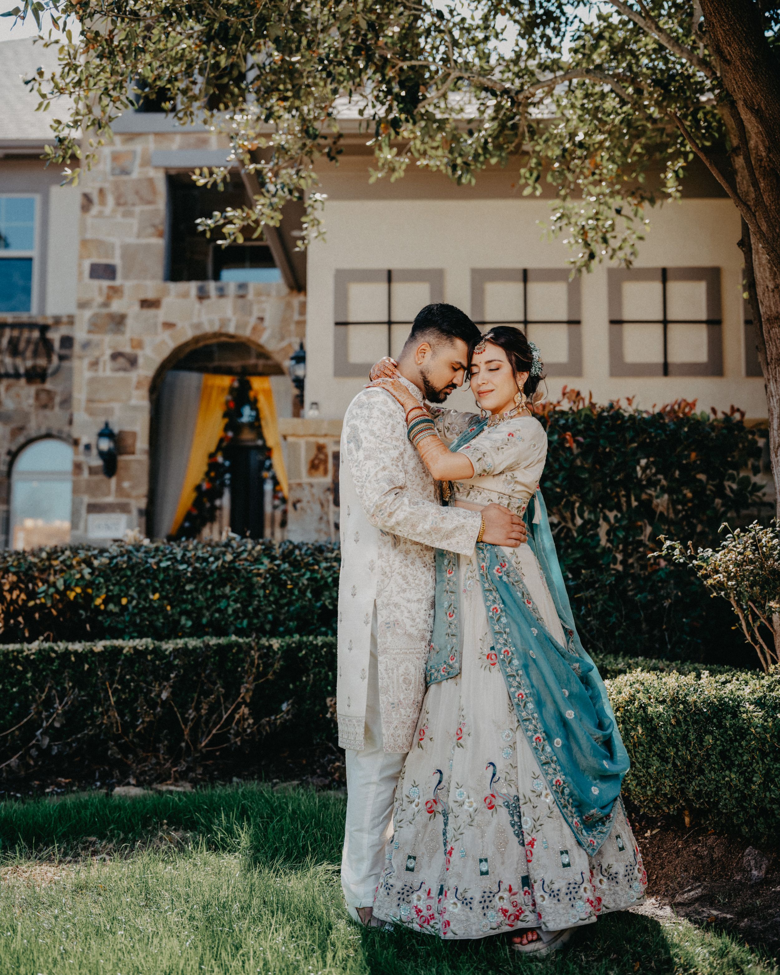 A romantic couple in traditional dress share tender moments in a garden setting under tree branches.