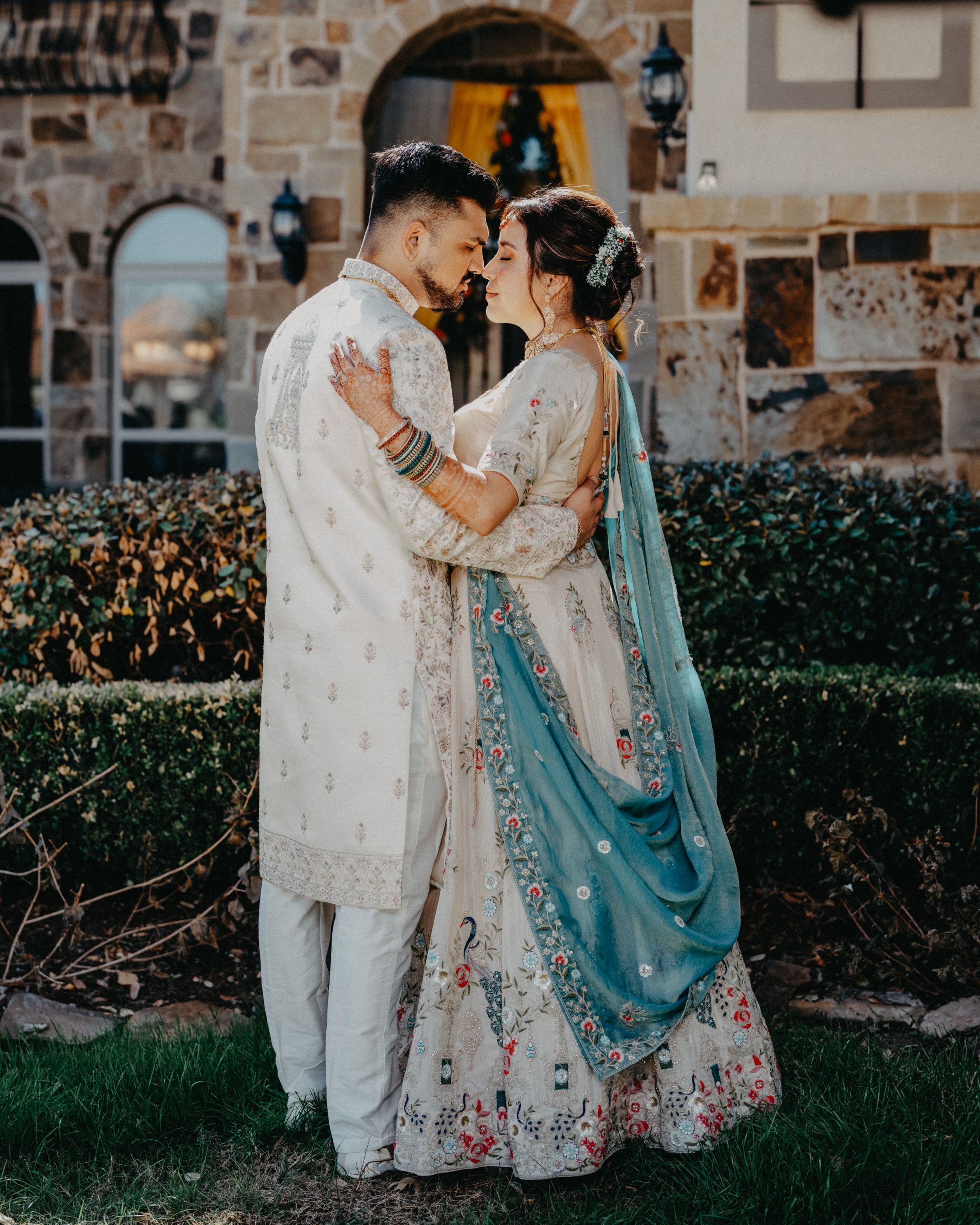 Couple in traditional South Asian wedding attire embrace outdoors against stone wall with turquoise dupatta flowing.