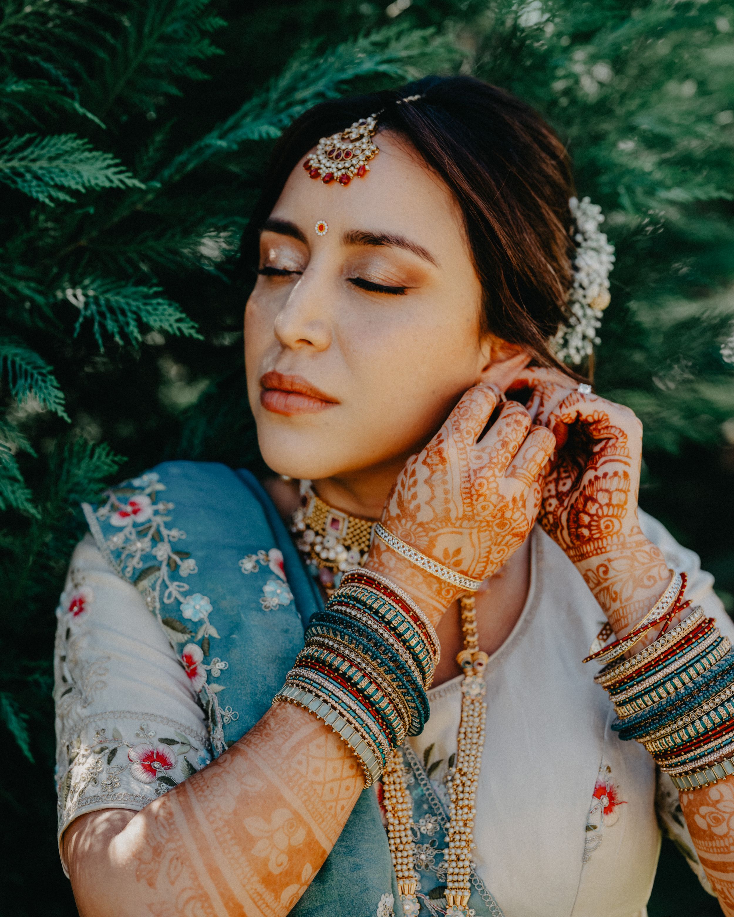 Close-up portrait showing intricate henna designs on hands and traditional Indian bridal jewelry against a green backdrop.