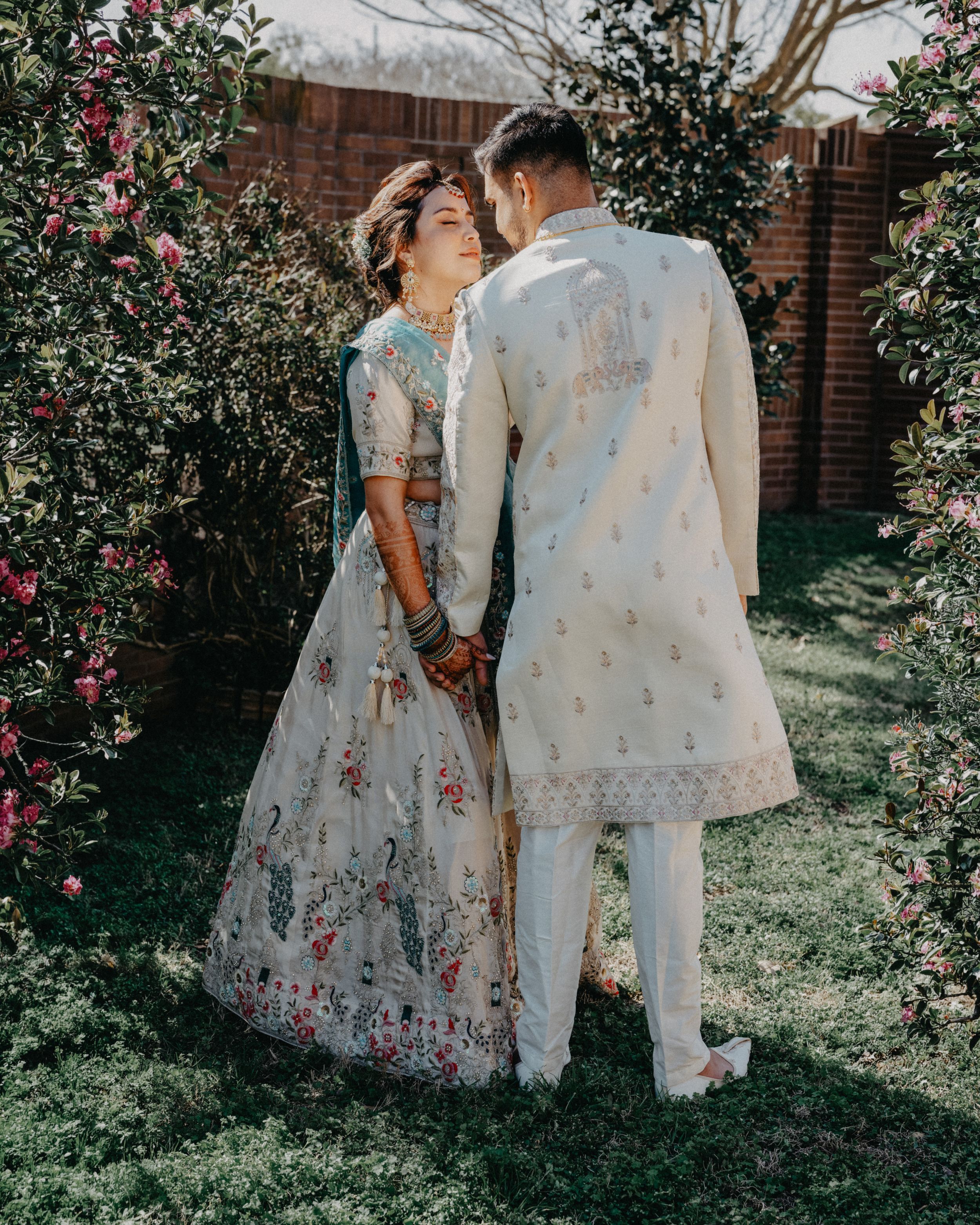A couple in traditional South Asian attire poses together in a garden with blooming flowers and greenery.