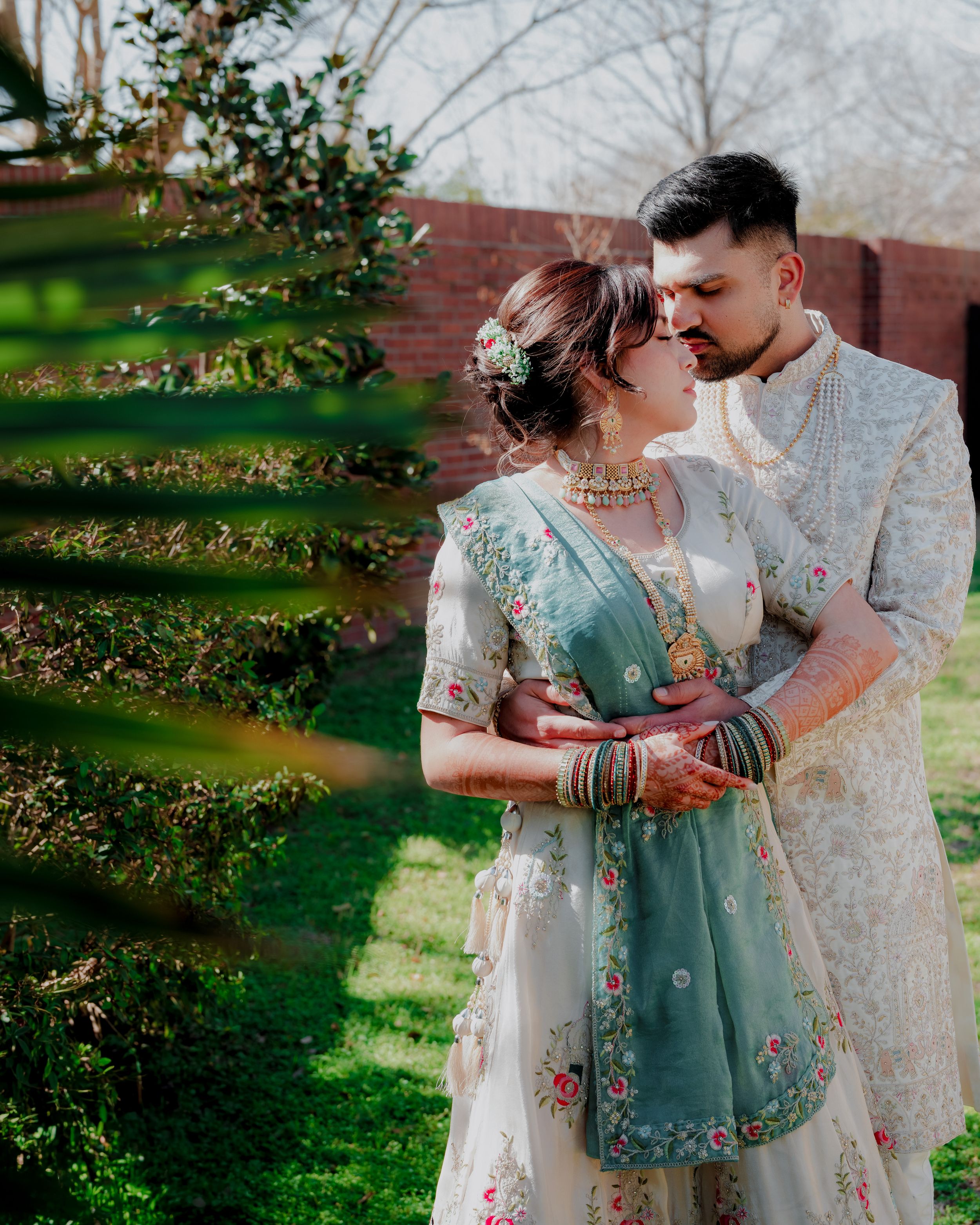 A couple in traditional South Asian wedding attire embraces in a garden setting with spring blossoms.